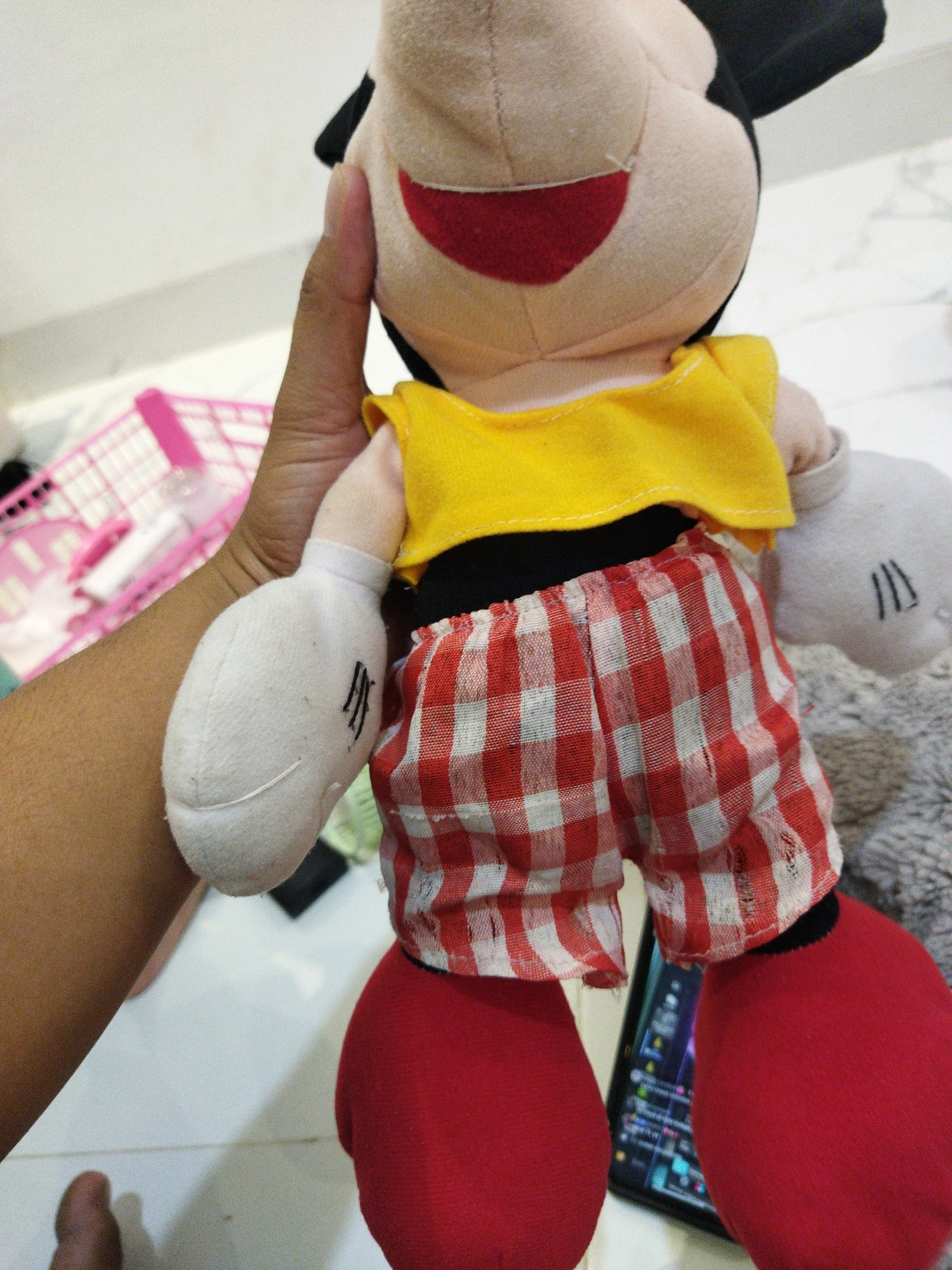 A close-up, eye-level, indoor shot captures a person's hand holding a Mickey Mouse plush toy. The plush toy, predominantly tan with red and white checkered shorts and red shoes, is the central subject. The background reveals a tiled floor, a pink plastic basket, and a grey fuzzy surface. The image is taken in Banjar Agung, Indonesia.