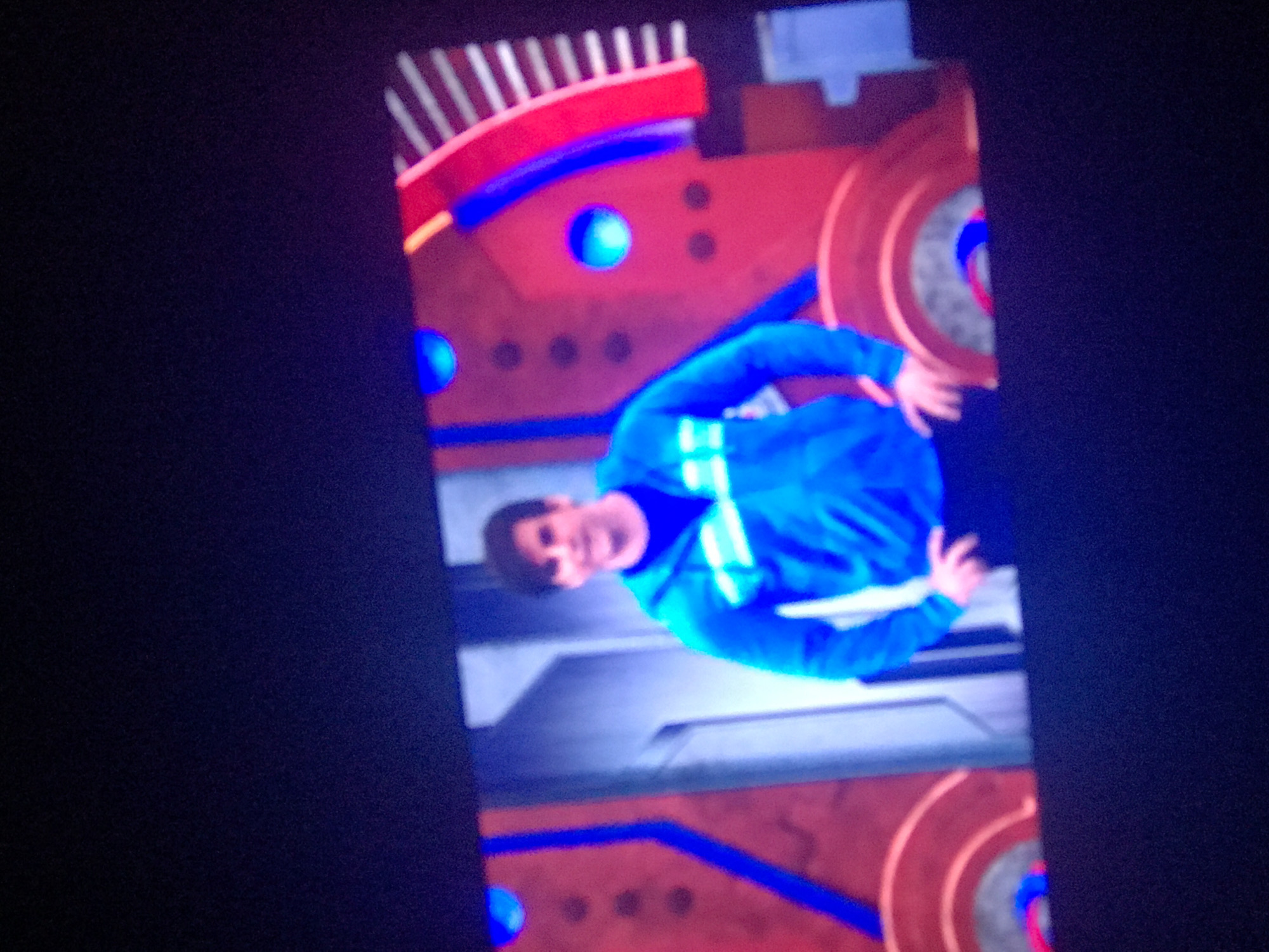 The image shows a man in a blue shirt with white stripes, standing in front of a brightly colored backdrop. The backdrop is primarily red and blue with circular elements. It is difficult to ascertain any location. Yerevan, Armenia is not identifiable in the image.