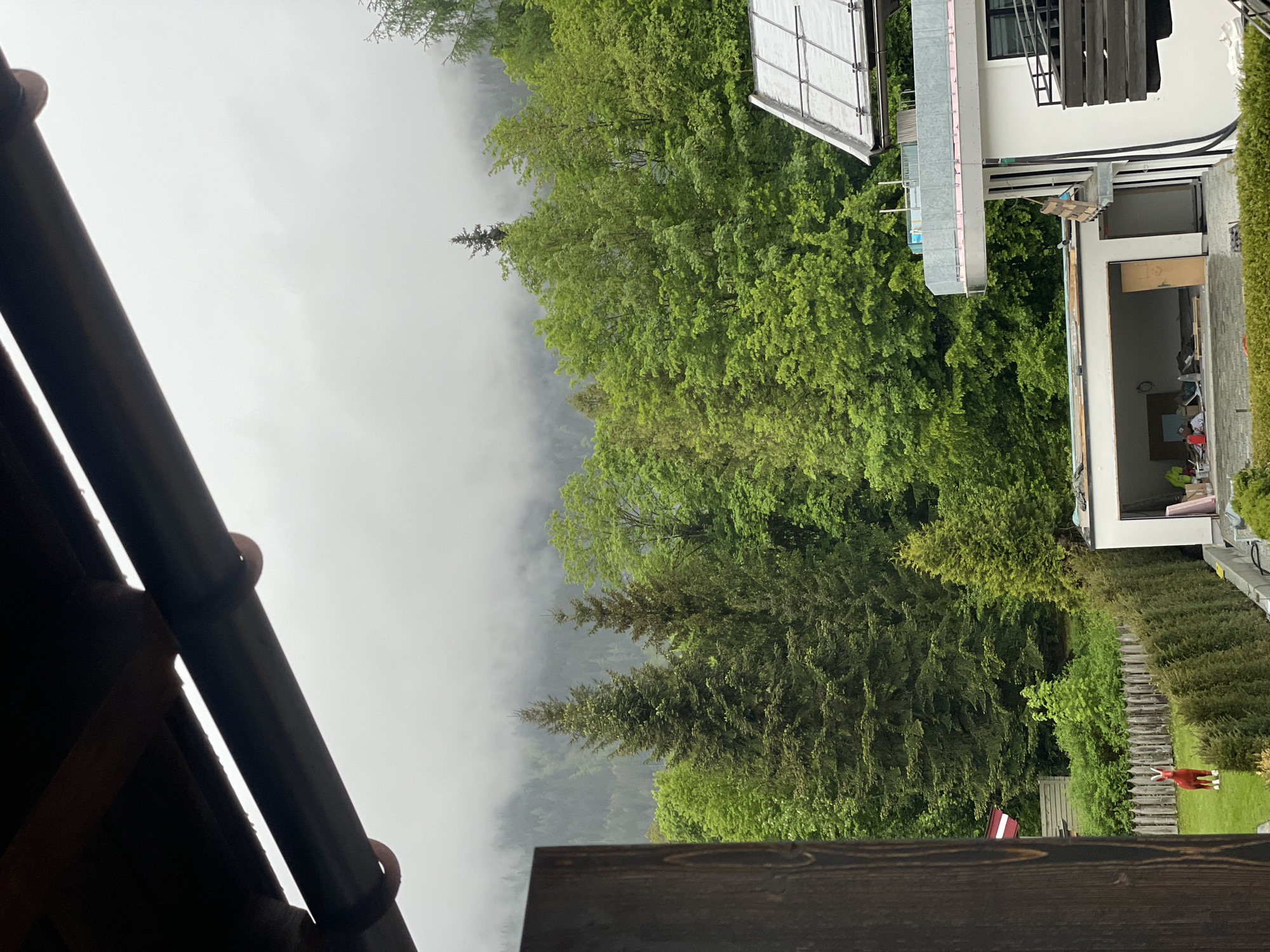 The image shows an outdoor scene, apparently from a residential area in Unknown, Unknown. The view is partially obscured by elements of the house the photo was taken from, including a brown roof and a wooden railing. Beyond the house, there's a dense area of green trees and what appears to be a misty mountain landscape in the background. To the right, there's a partial view of another house under construction, with visible building materials and an open doorway. In the foreground, a small green lawn is visible with a red plastic reindeer figure.