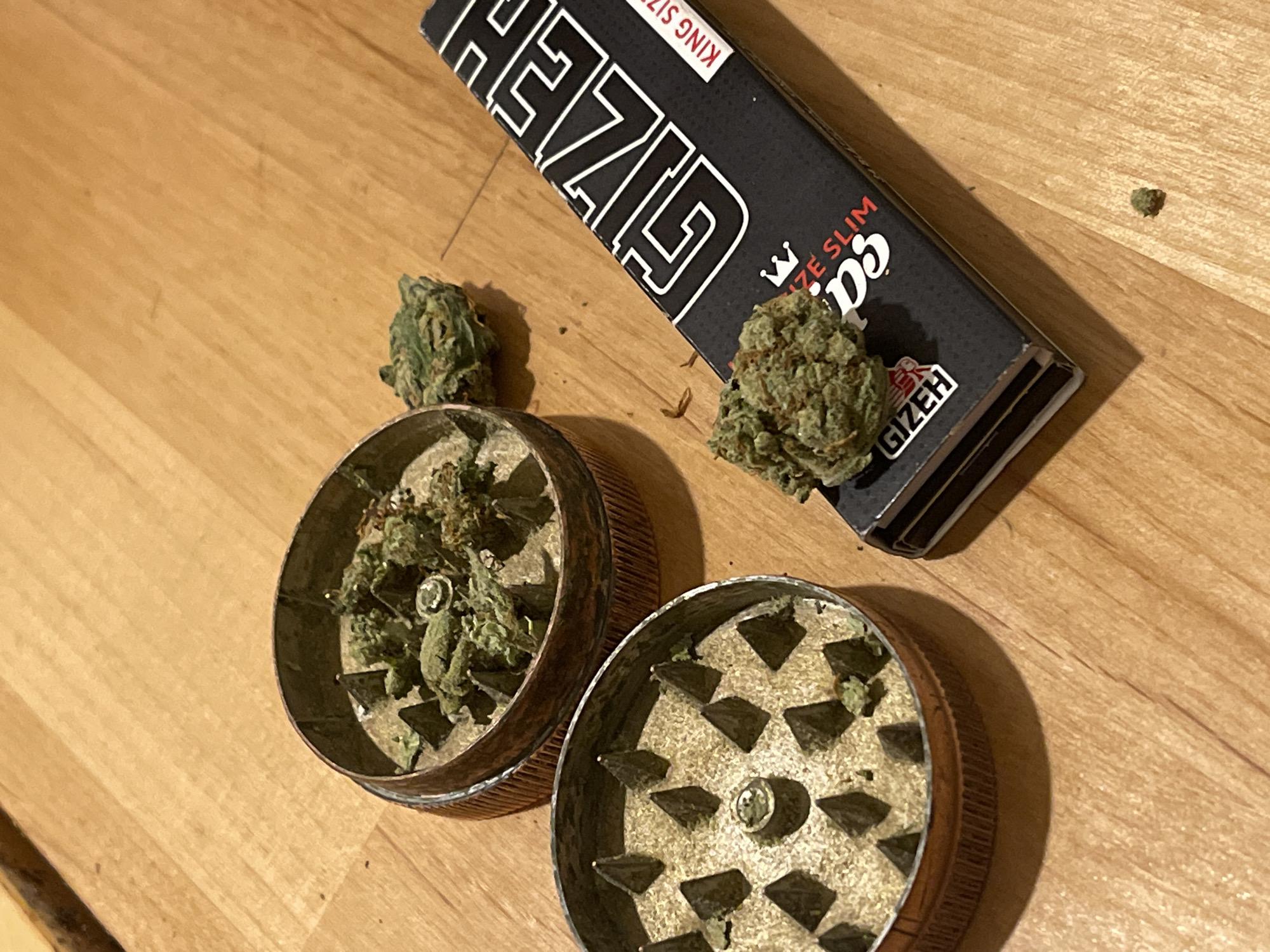 A high-angle indoor shot depicts a collection of cannabis, a metallic grinder, and a packet of rolling papers arranged on a light-colored wooden surface. The scene is located in the city of Unknown, Unknown.
The grinder, appearing to be made of aged copper or brass, is open and separated into two halves. The top half, positioned in the lower-left quadrant, displays sharp, conical grinding teeth arranged radially around a central screw. Small fragments of green plant matter are visible clinging to its surface. The bottom half, situated centrally and slightly behind the top half, contains a substantial amount of partially ground green cannabis buds within its grinding chamber, which also features similar conical teeth.
Multiple distinct, compact green cannabis buds are scattered across the scene. A prominent bud rests on a black packet of "GIZEH KING SIZE SLIM" rolling papers, which is positioned on the right side of the frame. The rolling paper packet has white text and a small red crown logo. Another smaller bud is visible between the two separated grinder halves. A tiny, individual fragment of cannabis lies on the wooden surface towards the bottom right corner of the image.
The underlying surface is a clean, polished light-brown wooden plank or flooring, exhibiting distinct wood grain patterns. The lighting is even and warm, originating from an unseen source, casting minimal shadows. No individuals or active interactions are depicted in the static arrangement.