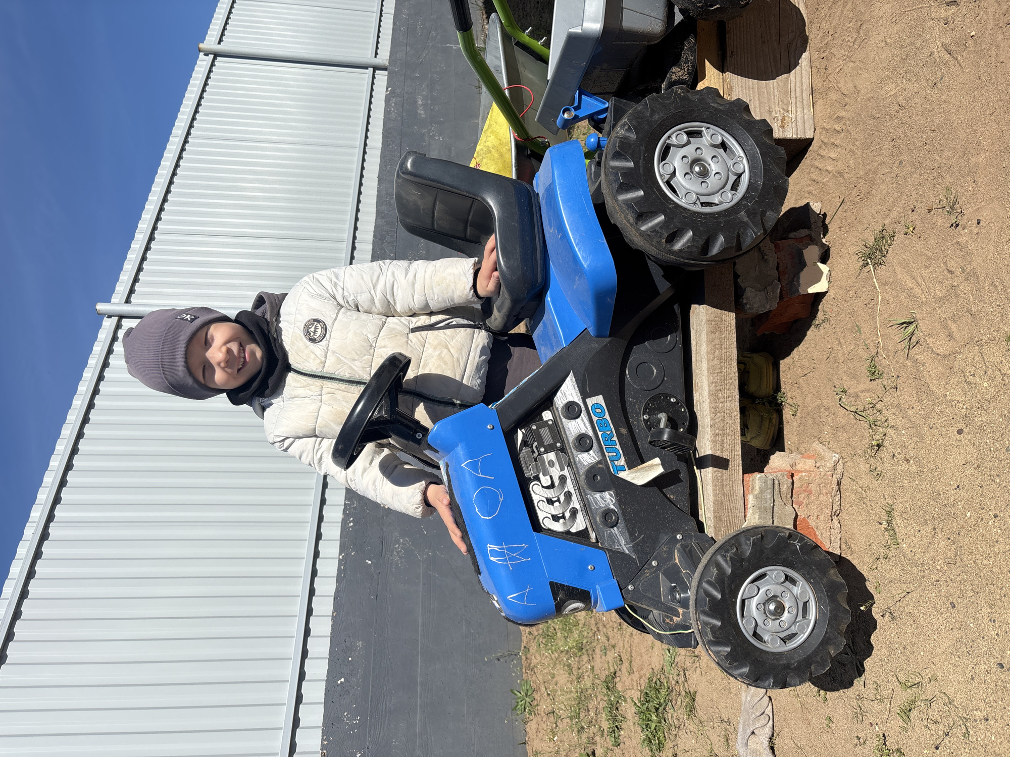 A child is sitting on a blue and black toy tractor outside in Chaikovsky, Russia. The child, wearing a white quilted jacket and gray beanie, is smiling. The tractor is partially elevated by wooden planks and bricks. The ground is sandy and grassy. In the background, there is a gray metal fence and a clear blue sky.