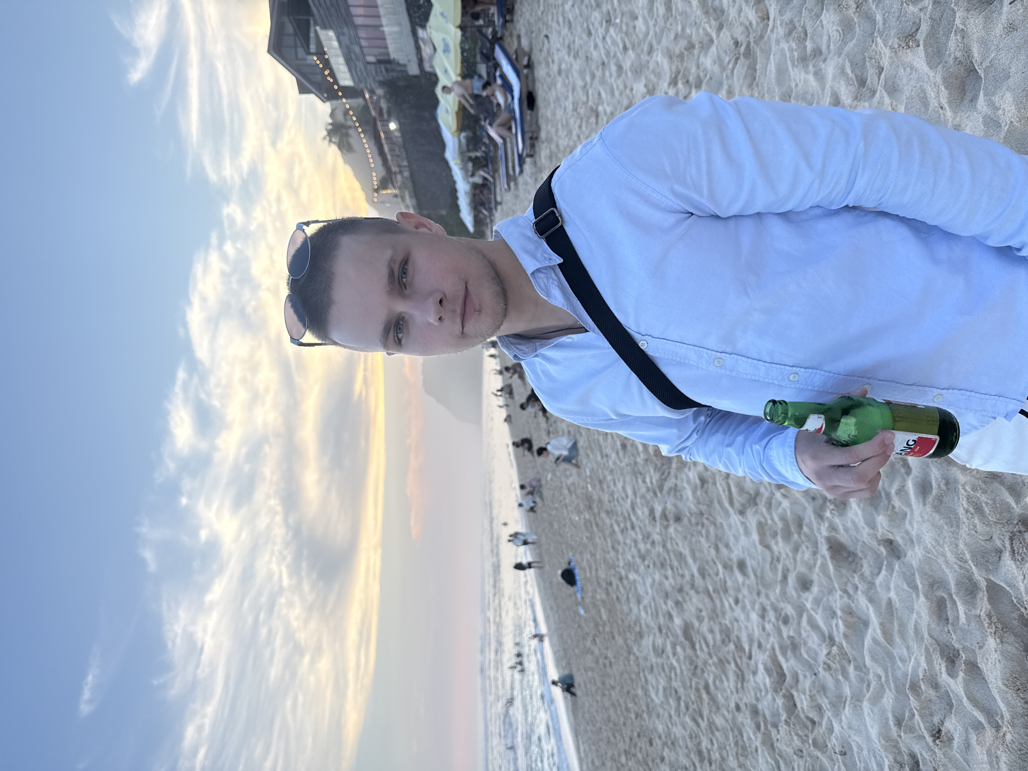 A man stands on a sandy beach in Kuta Selatan, Indonesia, holding a green beer bottle. He is wearing a light blue button-down shirt, sunglasses perched on his head, and a black shoulder strap across his chest. The beach has numerous people, and some are sitting on lounge chairs with yellow umbrellas. A building is in the background with lights. The sky is a mix of blue, white, and golden hues from the setting sun.