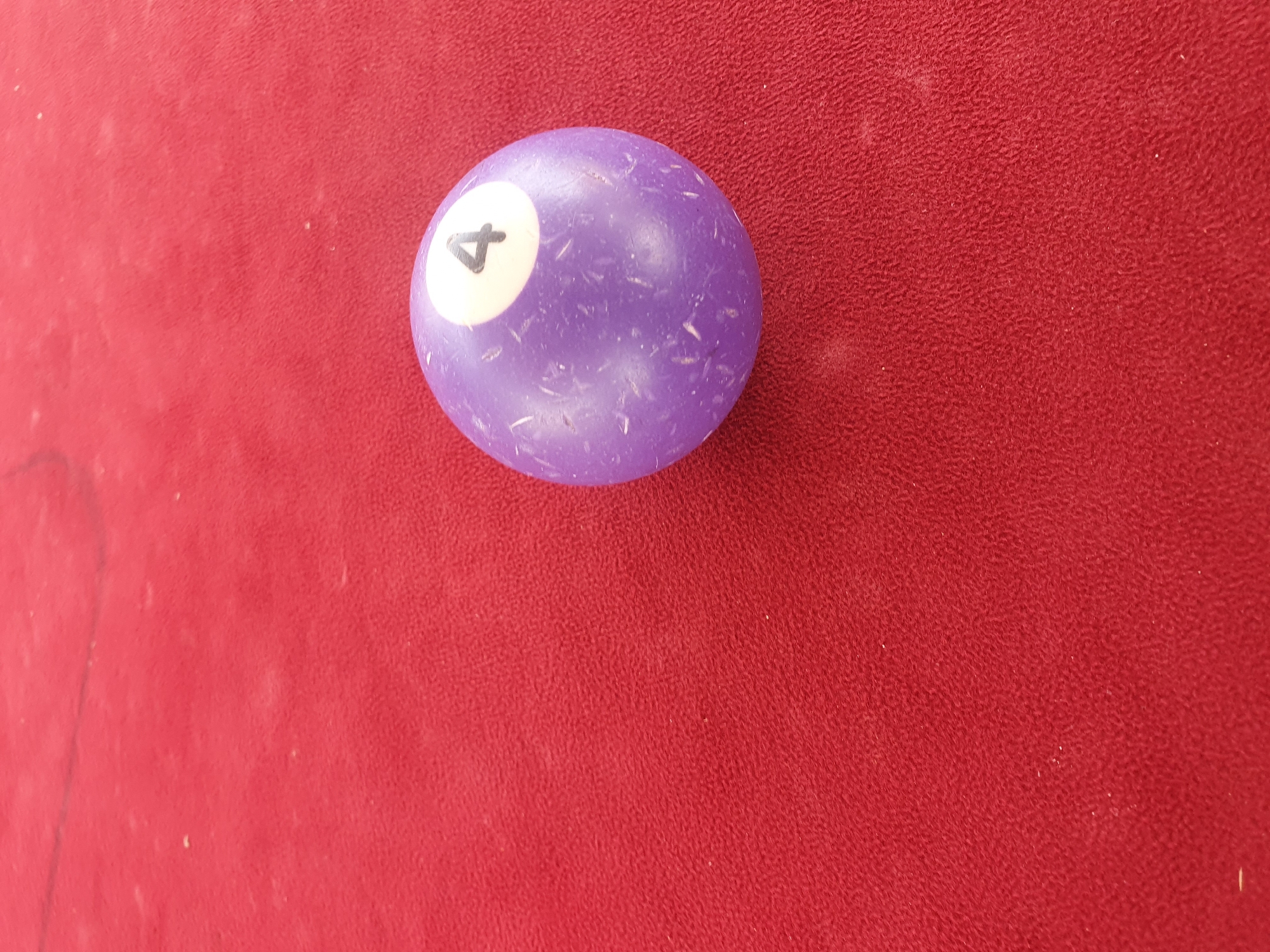 A single purple billiard ball with the number 4 printed in white is positioned on a red felt surface. The ball is slightly off-center, towards the right side of the frame. The red surface appears to be that of a billiard table, with a textured, felt-like appearance. There are some faint white specks visible on the surface of the ball and the felt, possibly dust or chalk residue. The lighting suggests an indoor environment, and the overall impression is a close-up shot of a billiard ball on a table.