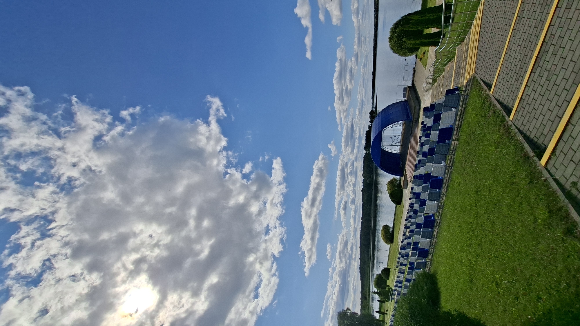 The image shows an outdoor amphitheater facing a body of water, likely a lake or reservoir, in the city of Myadzyel, Belarus. The amphitheater consists of rows of blue and white chairs arranged on a grassy slope. In the background, a stage is covered by a large blue arched structure. The water stretches to the horizon, where it meets a treeline and the sky. The sky is a vibrant blue with scattered white and gray clouds, and the sun is partially visible through some of the clouds, casting a bright light. The foreground features a paved walkway with yellow stripes, leading up to the seating area. The overall scene suggests a public gathering space designed for outdoor performances or events, set within a natural landscape.