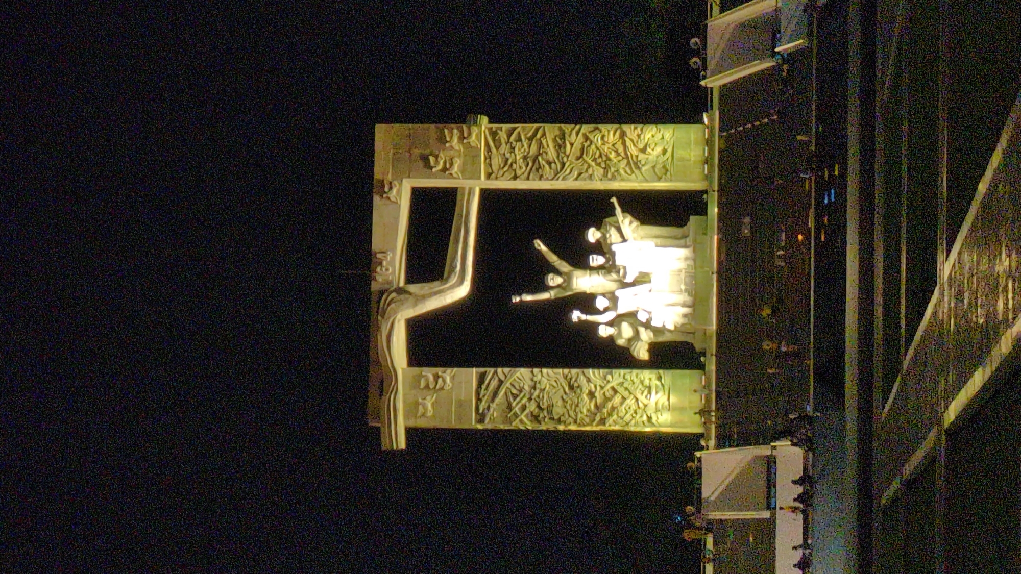 A dimly lit nighttime shot presents a monument under illumination, seemingly in Phan Rang – Tháp Chàm, Vietnam, given the location. The monument features a large sculptural frame, likely of carved stone, housing a multi-figure statuary grouping within. The statuary is lit strongly, emphasizing the figures. The frame itself appears to have bas-relief details along its top and bottom portions. The monument is situated adjacent to an elevated structure with railings. The surrounding environment is dark.