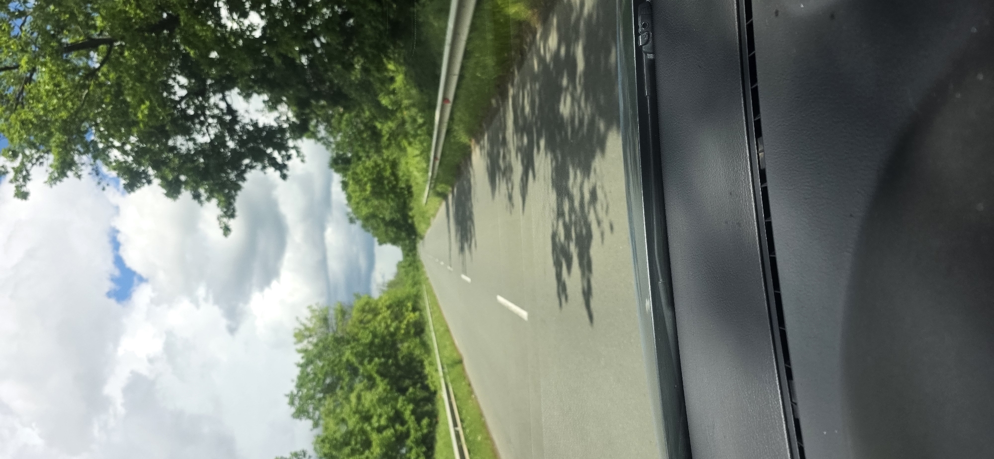 The image shows a view from inside a vehicle, likely a car, driving on a road. The road is surrounded by green trees and vegetation on both sides. There are white clouds and some blue sky visible above the trees. The road has white lane markings. There is a guardrail visible along the side of the road. Part of the vehicle's interior is visible on the right side of the image. The location is not evident from the image, and therefore Dragantsi, Bulgaria cannot be confirmed.