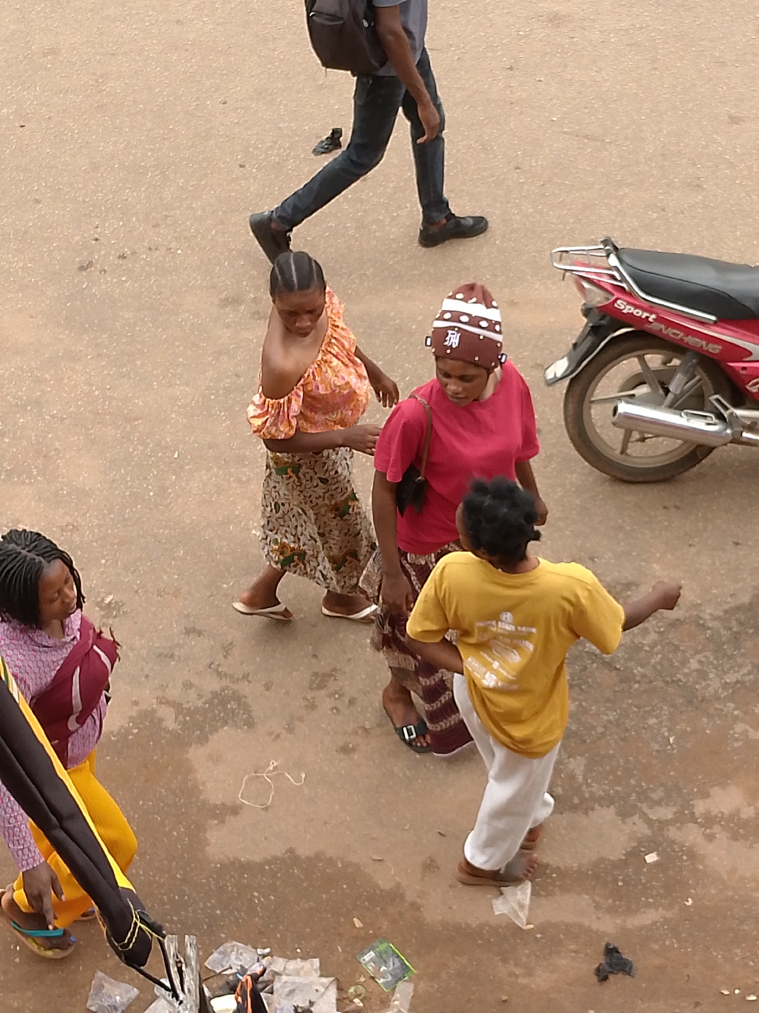 An overhead medium shot shows five people and a red motorcycle on a dusty, light brown asphalt surface in what appears to be an outdoor marketplace or street. On the left, a person in a pink and maroon patterned top and yellow pants is partially visible, wearing a yellow sandal. Next to them, a woman with dark braided hair wears a maroon headscarf and a purple and pink patterned top. To her right, a woman in an orange and pink patterned off-the-shoulder top and a patterned skirt walks away from the viewer. In the center, a woman wearing a pink t-shirt and a maroon and white patterned hat converses with a young person wearing a yellow t-shirt and white pants. The young person is gesturing with their right hand. A red motorcycle with a silver exhaust pipe is parked on the right side of the frame. The ground is littered with small pieces of debris, including plastic bags and paper. In the background, the lower legs and feet of a person wearing dark pants and black shoes are visible, walking away from the scene.