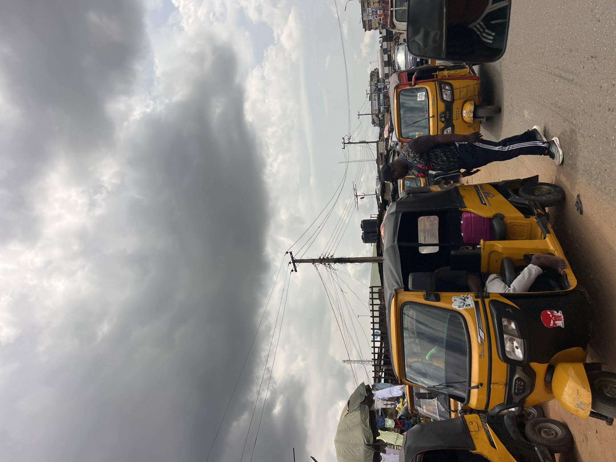 The image depicts a street scene in Nkpor, Nigeria, characterized by a traffic jam of yellow auto-rickshaws, commonly known as "keke." The sky is overcast with dramatic clouds, suggesting a cloudy or potentially rainy day. The perspective is from within one of the keke, looking forward, with a side mirror reflecting a fragment of another vehicle and a glimpse of the sky.
In the foreground, a man wearing a black cap, a patterned t-shirt, and dark tracksuit pants with white stripes is leaning against a keke, with his arms extended. Another keke is partially visible behind him. The keke are parked closely together, indicating heavy traffic. In the middle ground, more keke are lined up, with some passengers visible inside. The street is paved and dusty. In the background, buildings and various structures line the street, including what appear to be shops and stalls. Power lines and utility poles crisscross the scene, common in urban and semi-urban areas. Some laundry is hanging on lines between buildings in the distance.
The overall impression is one of a busy, congested street in Nigeria during the day, under a cloudy sky. The activity is primarily people waiting in traffic in their vehicles. There are no obvious signs of urgency or distress, suggesting this is a common occurrence. Some decals are visible on the yellow keke, including what appears to be a cartoonish character and a logo with text. One keke has "TVS" written on its front. Another keke has "DANKO" written on its side.