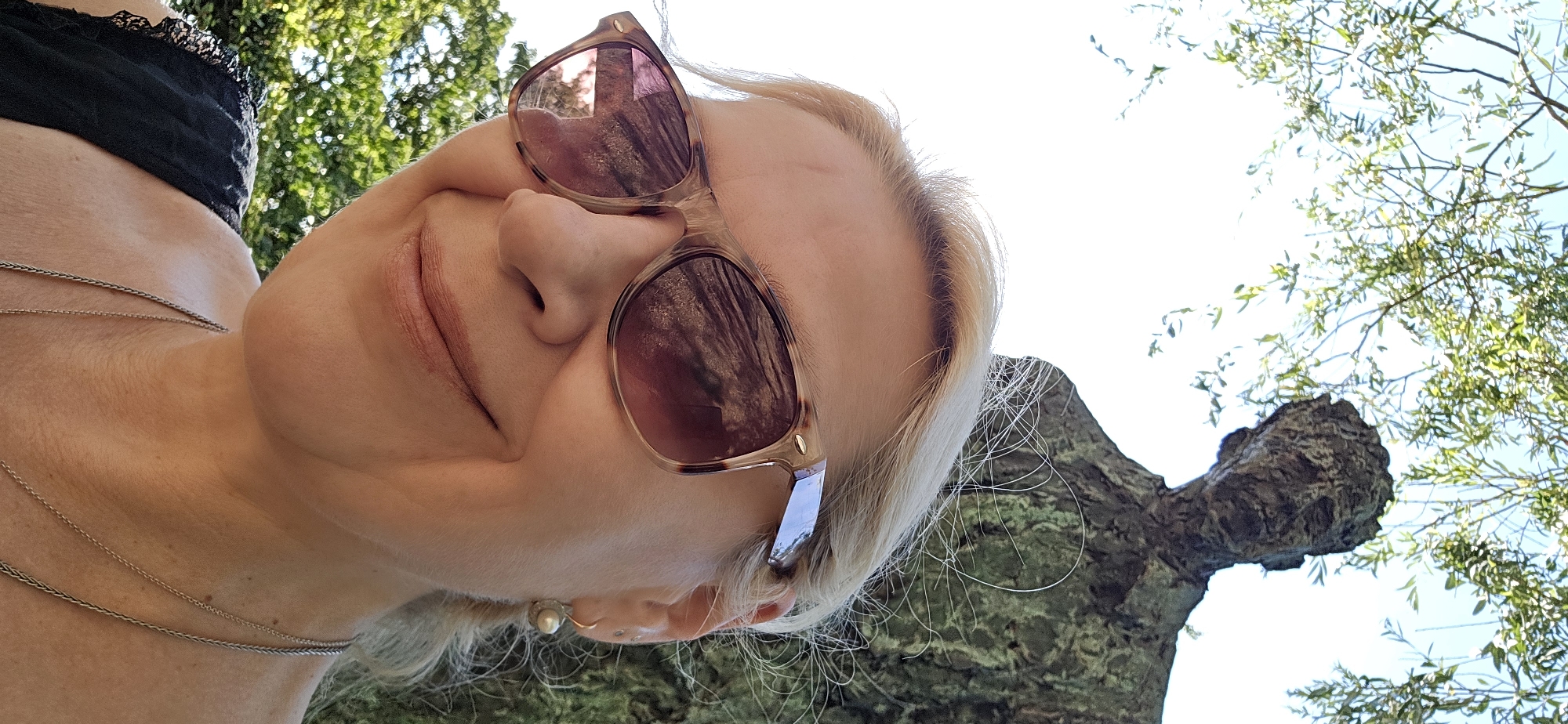 A medium close-up selfie shows a smiling woman from the chest up, positioned in the foreground. She has blonde hair and is wearing tortoiseshell sunglasses and a pearl earring. Behind her, out of focus, is a textured, weathered statue of a man's head and shoulders, possibly bronze, with a large tree branch and green foliage overhead against a bright sky. The overall impression is that the photo was taken outdoors in a park or public space, possibly a sculpture garden, with dappled sunlight filtering through the leaves. The statue's rough, moss-covered texture suggests it is old and has been exposed to the elements. The woman's attire and relaxed demeanor imply a casual outing.