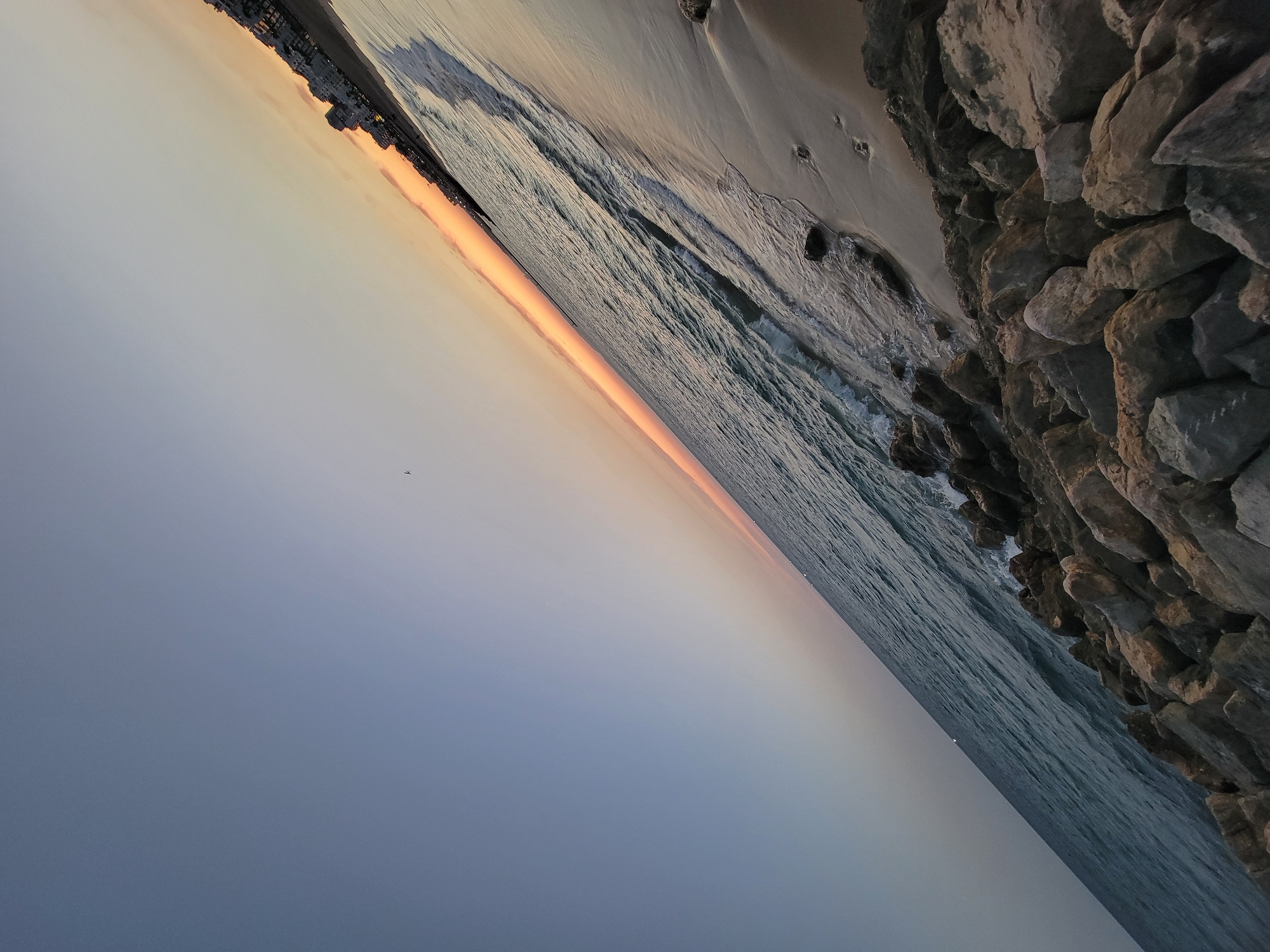 This is a photograph of a beach in Quarteira, Portugal, taken at an angle. The sky is clear and gradually transitions from a pale blue at the top to a pale orange at the horizon line. A thin band of orange is visible along the horizon, indicating the presence of a sunset or sunrise. The ocean occupies the left side of the frame, with gentle waves lapping at the shore. The water appears calm and a deep blue-green. The beach is sandy and runs along the right side of the image, meeting the ocean. Scattered on the sand are a few footprints. In the foreground and along the right edge of the image, a pile of large, rough rocks forms a breakwater or part of the coastline. In the distance, along the horizon, a line of buildings characteristic of a coastal town is visible. A single small bird can be seen in the sky. The overall scene depicts a serene coastal landscape at dusk or dawn.