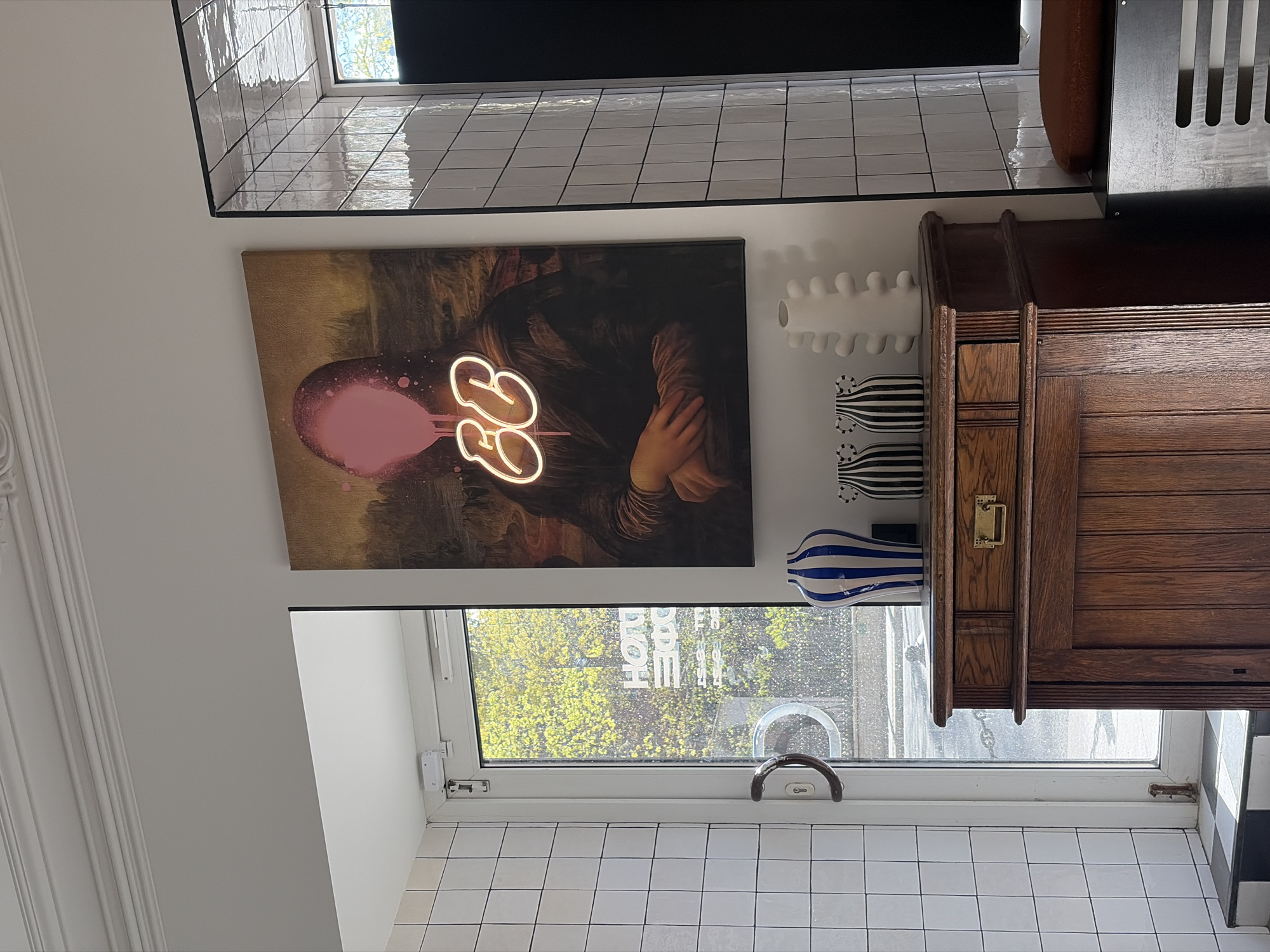 A kitchen interior. A painting of a figure with a neon pink head and number symbols hangs on the wall. The wall is tiled white. A window looks out on trees and the reflection of a building. A cabinet is to the right of the painting. Above the painting, the wall is tiled in grey.
