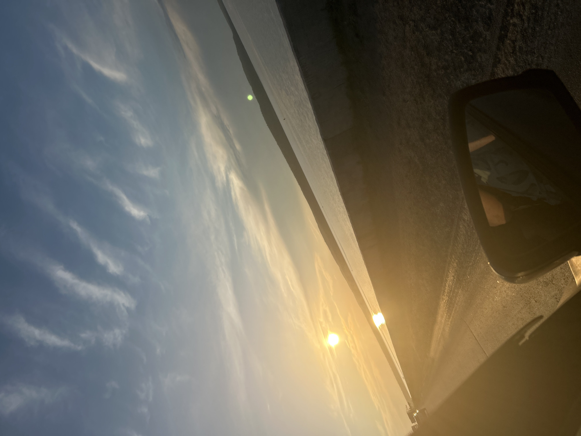 This is a wide-angle, slightly tilted, outdoor, eye-level shot of a sunset over a body of water. The sun, a bright orange orb, is low on the horizon, casting a golden glare across the scene. Wispy white clouds are streaked across a pale blue sky. A dark, sloping embankment runs along the edge of the water, and a paved road runs parallel to the embankment on the left side of the frame. The road is mostly in shadow, with a few vehicles visible in the distance. In the foreground, a car's side mirror is visible, reflecting a person wearing a light-colored shirt with a pattern. The image was taken from inside a vehicle, likely while driving. This scene is likely depicting the Kama River embankment in the city of Chaikovsky, Russia, given the presence of a large body of water and a road adjacent to it, common features of that location.