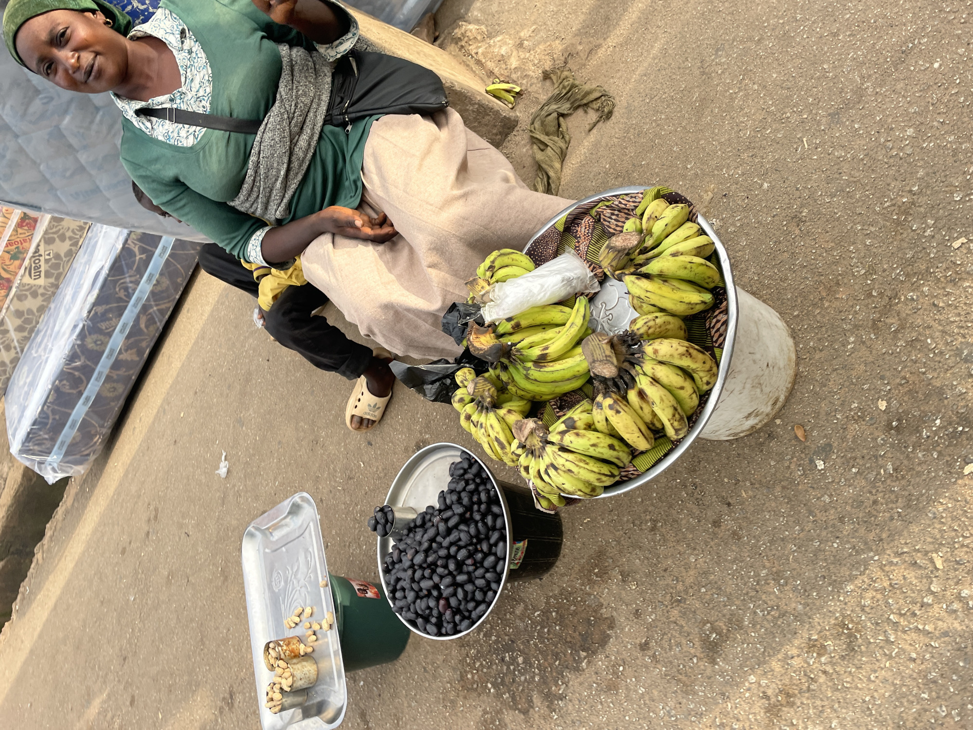 An overhead shot captures a woman sitting by a market stall in Jos, Nigeria, offering a selection of produce. The woman, wearing a green headscarf and a green top with a patterned collar, is seated on a raised concrete block. She has a woven grey fabric wrapped around her waist and a dark bag slung over her shoulder. To her left, three large metal bowls contain fresh produce. The largest bowl, placed on a black bucket, is filled with dark, oval-shaped berries. Another metal bowl, positioned on a green bucket, holds a tray with small cans of nuts. A third, wider bowl, sitting on the ground, is filled with bunches of unripe green bananas. In the background, several mattresses are stacked and wrapped in plastic, suggesting a nearby shop or stall. The ground is paved with concrete, typical of a street market environment. A child, wearing a yellow shirt and black pants, is partially visible behind the woman. The overall scene depicts a vibrant street market with a vendor attending to her goods.