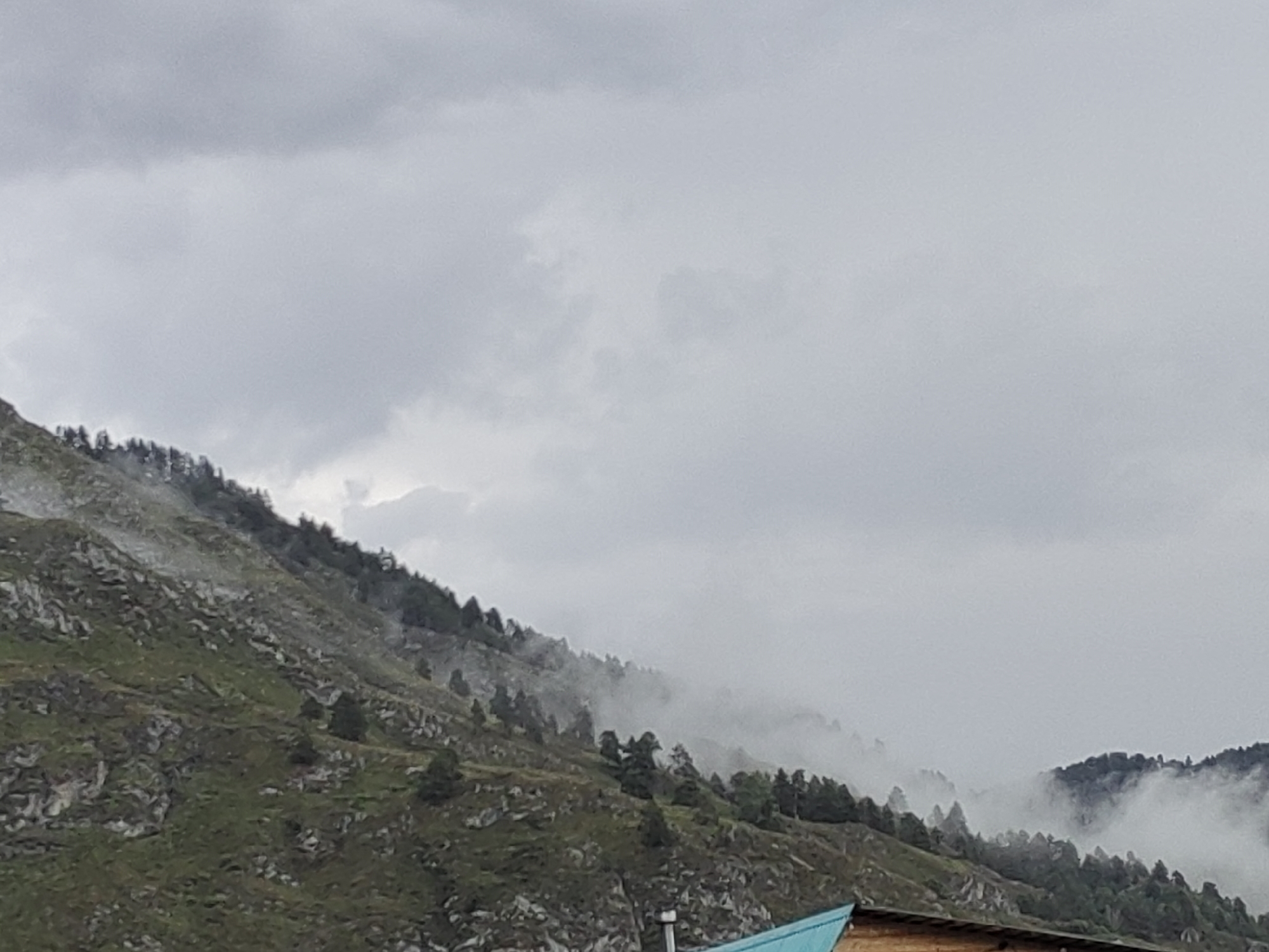 The image depicts a cloudy sky over a mountain landscape. The mountain slopes are covered with sparse vegetation and trees. Mist or fog is present on the mountains, obscuring parts of the scene. In the foreground, a portion of a wooden structure with a blue roof is visible. There are no people or animals in the frame. The location in the city of Anos, Russia is not discernible from the image itself.