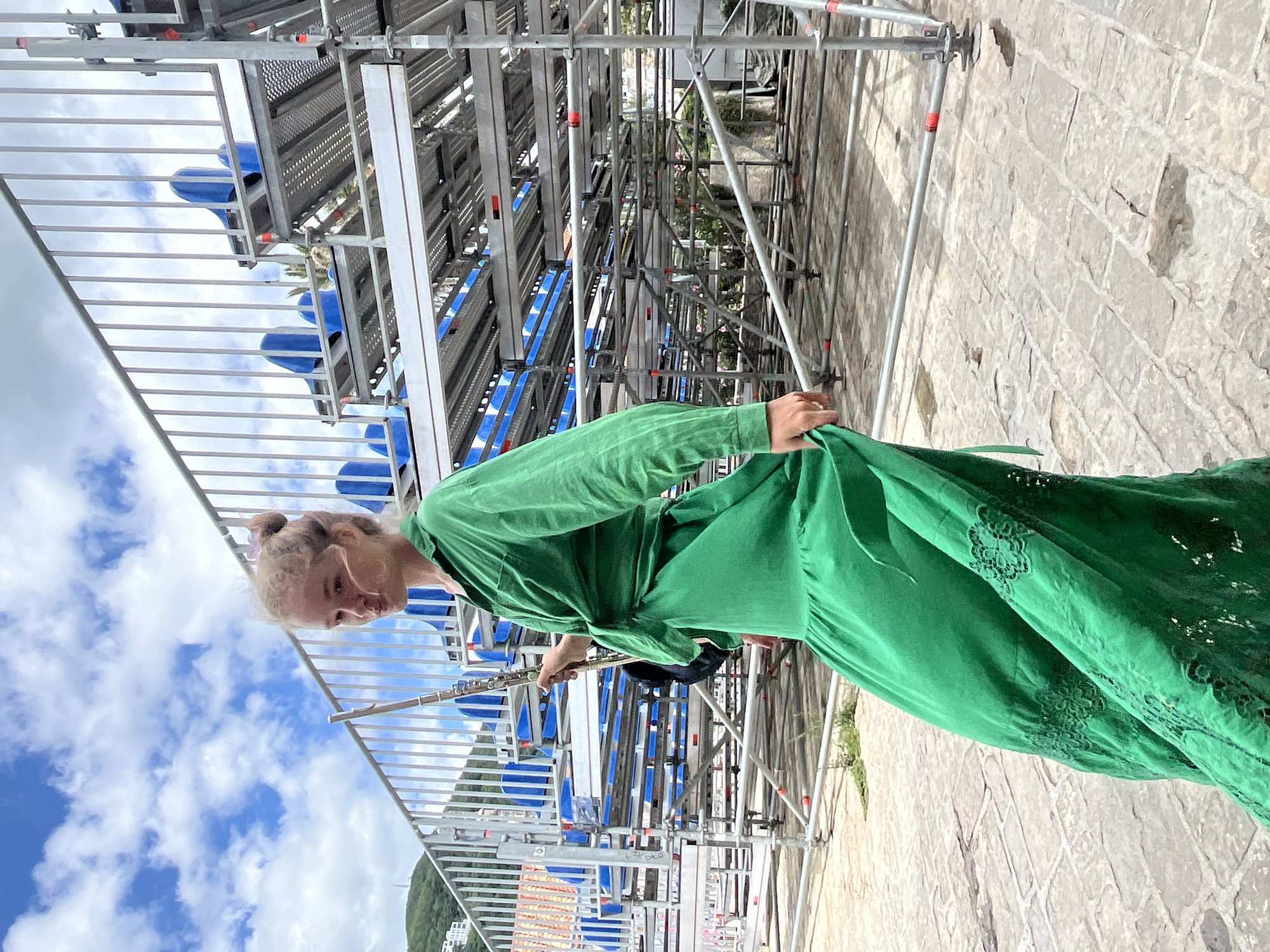 A young woman with blonde hair pulled into a bun walks past temporary scaffolding in Budva, Montenegro. She wears a long, flowing emerald green dress with lace details on the skirt and a tied knot at the waist. She is holding a flute in her right hand, with the instrument angled upwards. The scaffolding, likely for an event, is constructed of metal poles and platforms with blue seating visible in the background. The ground is paved with light-colored cobblestones, some of which appear wet or discolored. The sky above is blue with scattered white clouds. In the distance, a green, hilly landscape with buildings can be seen.