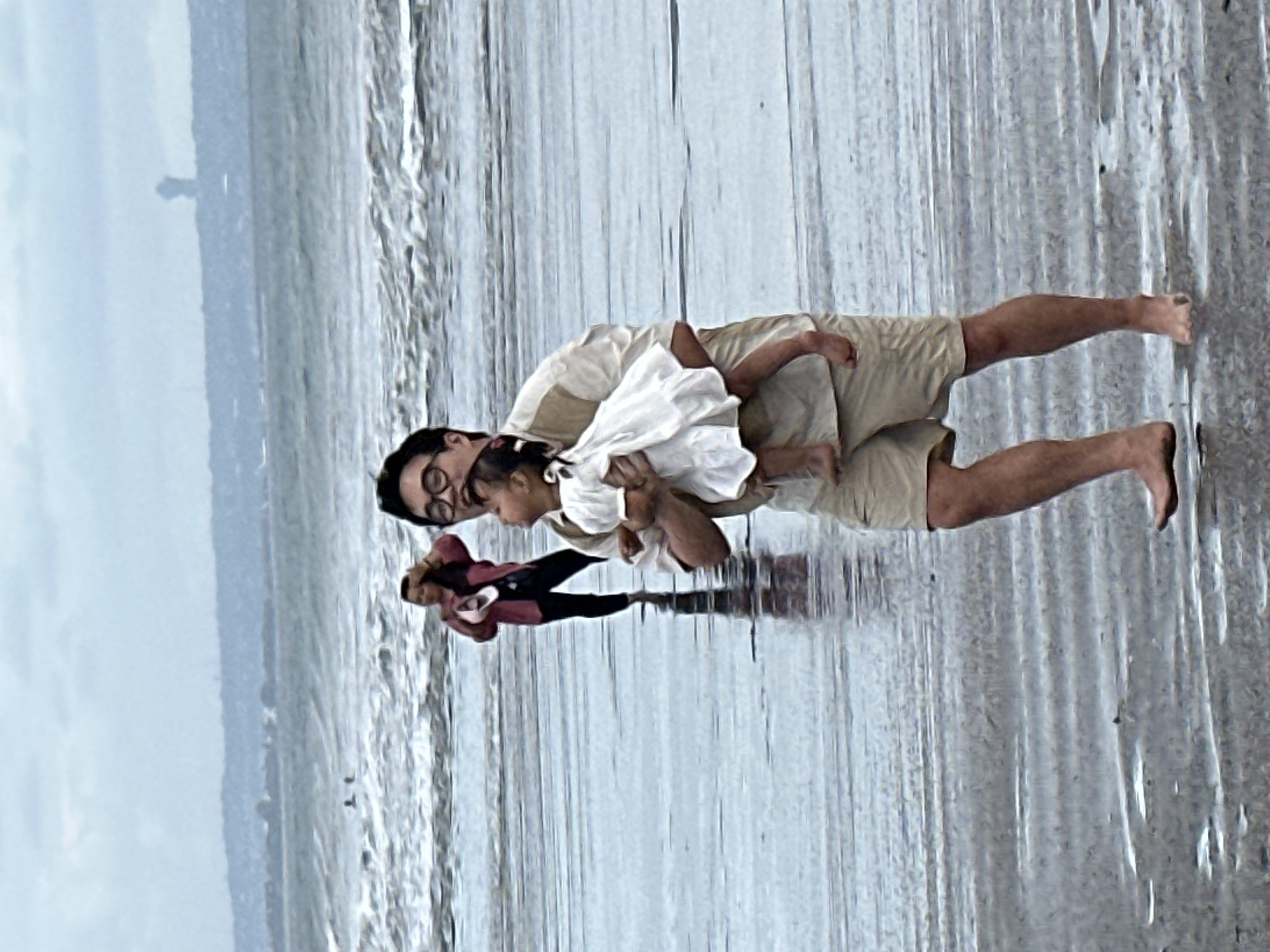 The image shows two people on a beach. A man is holding a young child in his arms. The man is wearing glasses, a light-colored shirt, and tan shorts. The child is wearing a white outfit. The background is a body of water and a sandy shore. The water appears to be shallow, with gentle waves. The sky is overcast, suggesting it may be a cloudy day. The overall scene appears peaceful and familial, as the man is embracing the child. There are no visible objects or text.