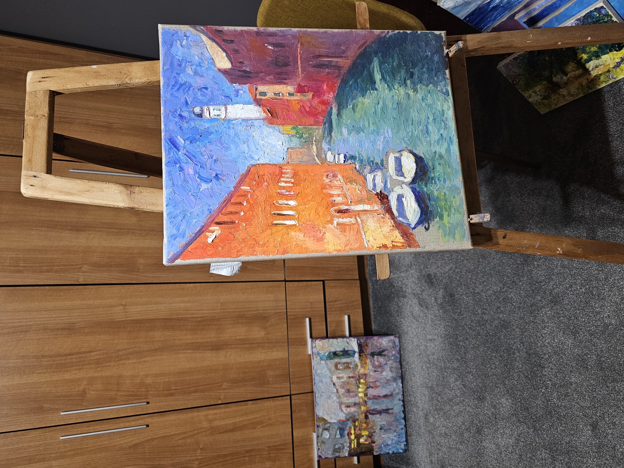 A painting is displayed on a wooden easel. The painting features an orange pathway or canal leading towards the center of the composition, flanked by blue and green hues on either side, potentially representing water or foliage. The upper portion of the painting has a building or structure rendered in shades of red and pink. The easel itself is made of light-colored wood, appearing simple and functional. In the background, there are closed wooden cabinets and a portion of another painting. The floor is covered in gray carpet. The setting appears to be indoors.