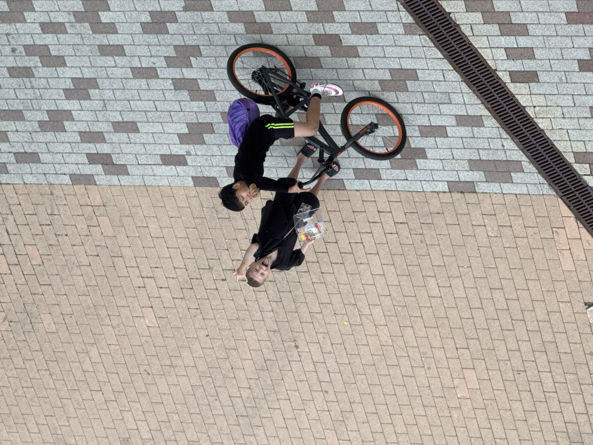 An aerial, full-shot image depicts two young boys outdoors on a paved walkway. The boy on the left, facing upwards, is wearing a black shirt and shorts, with a clear plastic bag containing colorful items. He is holding his right hand up in a "rock on" gesture and sticking his tongue out, seemingly playful. The boy on the right is seated on a black BMX bicycle with orange rims and is wearing a black long-sleeved shirt, black shorts with green stripes, and white and pink athletic shoes. He is holding the handlebars and looking forward. A purple backpack is attached to the back of the bicycle. The walkway consists of tan, grey, and dark grey rectangular paving stones. To the left of the boys, a black metal grate runs diagonally across the frame. The setting is a city street in Khimki, Russia.