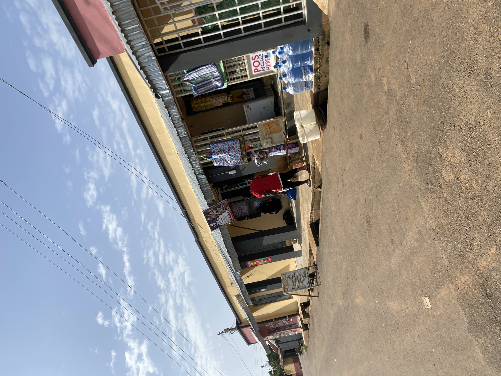 In Bar Jirgi Summa, Nigeria, an outdoor scene depicts a row of small shops with yellow and pink facades under a bright blue sky with scattered clouds. The shops feature corrugated metal awnings. In front of one shop, a person wearing a red shirt and black shorts is standing near a broom. Various items are displayed outside the shops, including shopping bags hanging from the awnings and posters advertising "POS AVAILABLE HERE." Several large blue water jugs are arranged on the ground in front of a gated window, next to a white bucket. A sign on an easel stands in front of another shop. The ground in the foreground is a paved road with some debris.