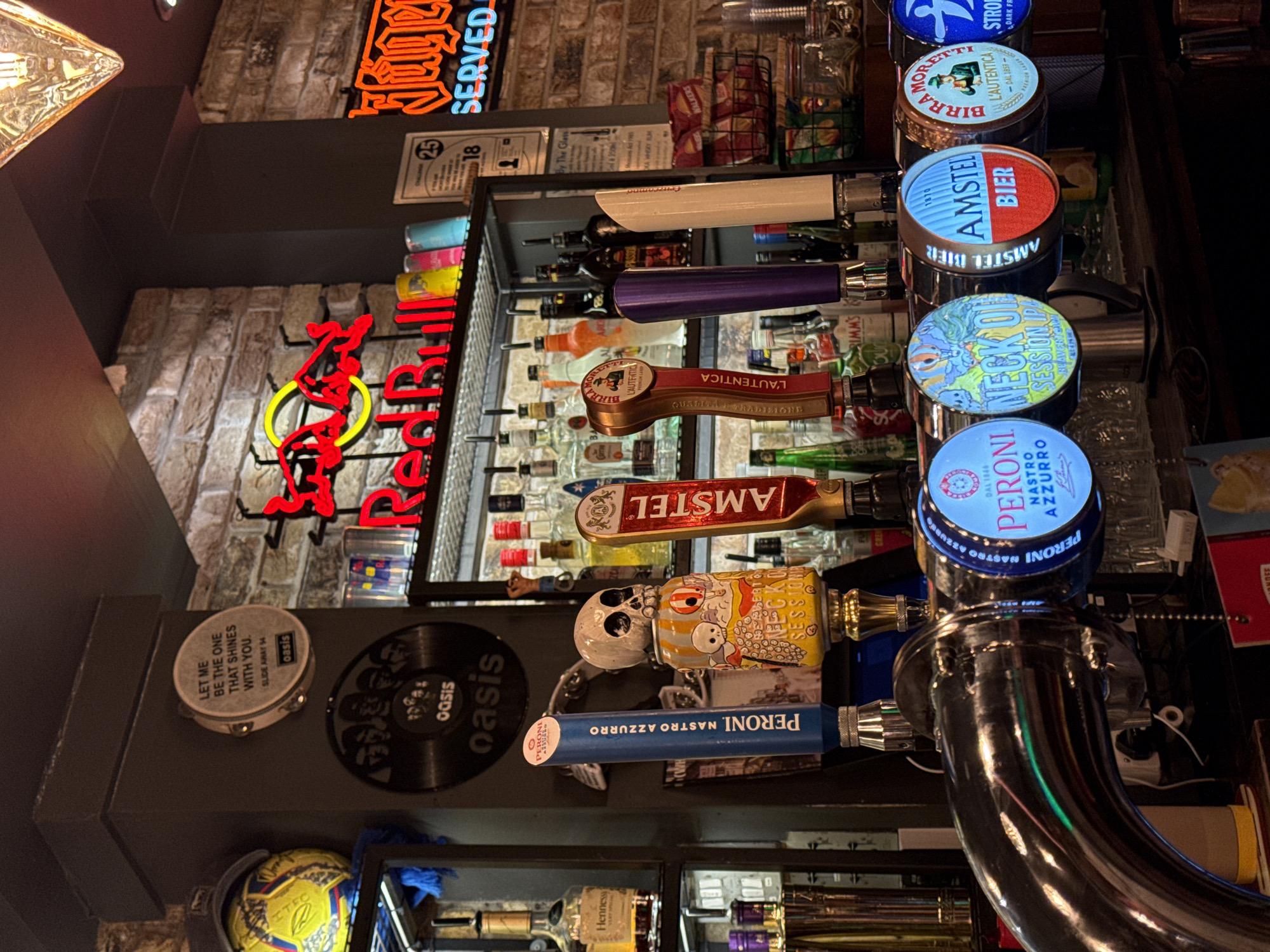This is a photograph taken inside a bar or pub in Ipswich, United Kingdom. The image focuses on a lineup of beer taps, featuring brands like Peroni, Neck Oil Session IPA, Amstel, Birra Moretti, and Peroni Nastro Azzurro. There are various beer tap handles that correspond to the different brands. Behind the taps, bottles of various alcoholic beverages are arranged on shelves. The background features brick walls with neon signs, including a Red Bull sign and a sign advertising food service. A few decorative items are present on the lower left, including a soccer ball and items labeled "oasis". The overall lighting in the location appears to be fairly dim.