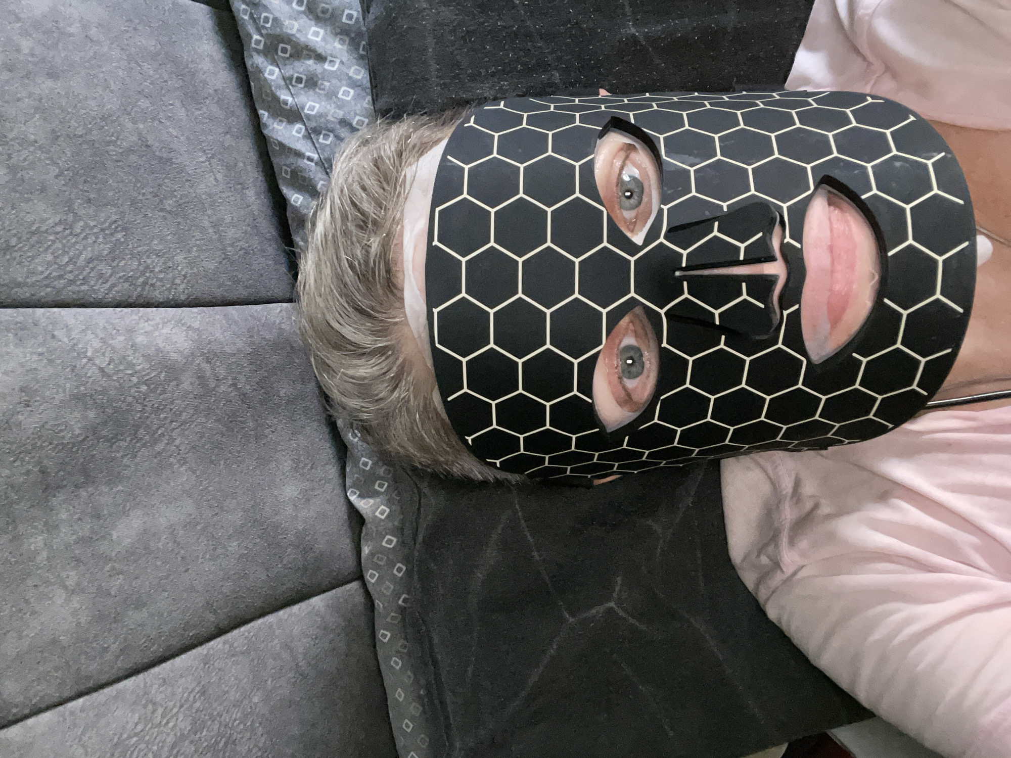 The image shows a person lying down, wearing a black and white hexagonal patterned mask that covers their face, exposing only their eyes and lips. The person has light-colored hair and is wearing a light pink shirt. They are lying on a dark gray surface, possibly a couch or bed, with a textured gray headboard visible behind them. The overall scene appears to be indoors. There is no discernible information in the image that would indicate a specific location in Kadıköy, Turkey.