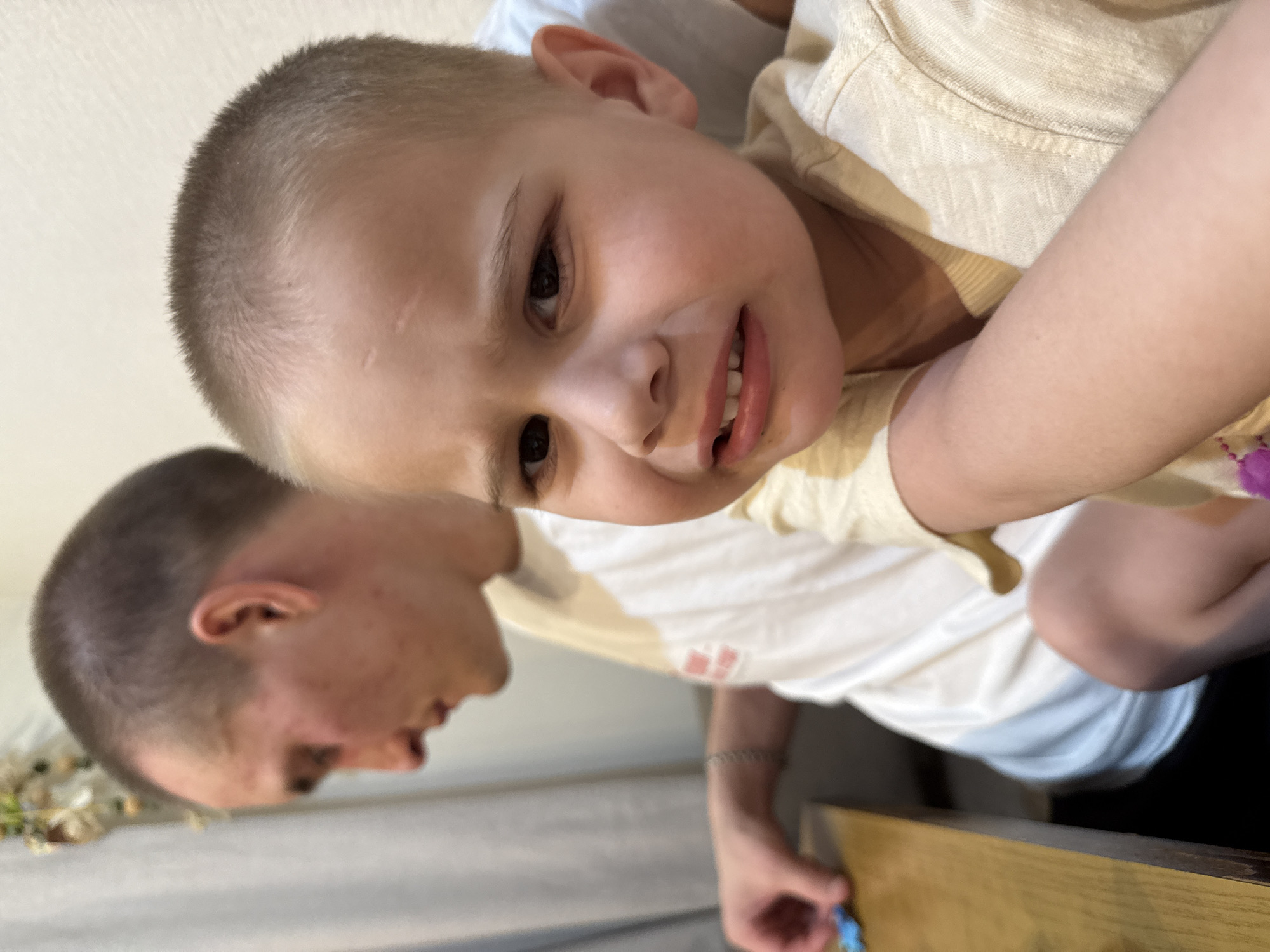 A close-up shot depicts a child with a buzz cut, wearing a beige t-shirt, and positioned in the arms of an individual with a similar haircut. The background is a white wall with a decorative floral piece on the left. Part of a wooden surface and a second person's arm are visible at the bottom of the frame. The location is likely indoors, based on the lighting and interior elements, but the city of Chaikovsky, Russia, cannot be confirmed based on the visual information provided.