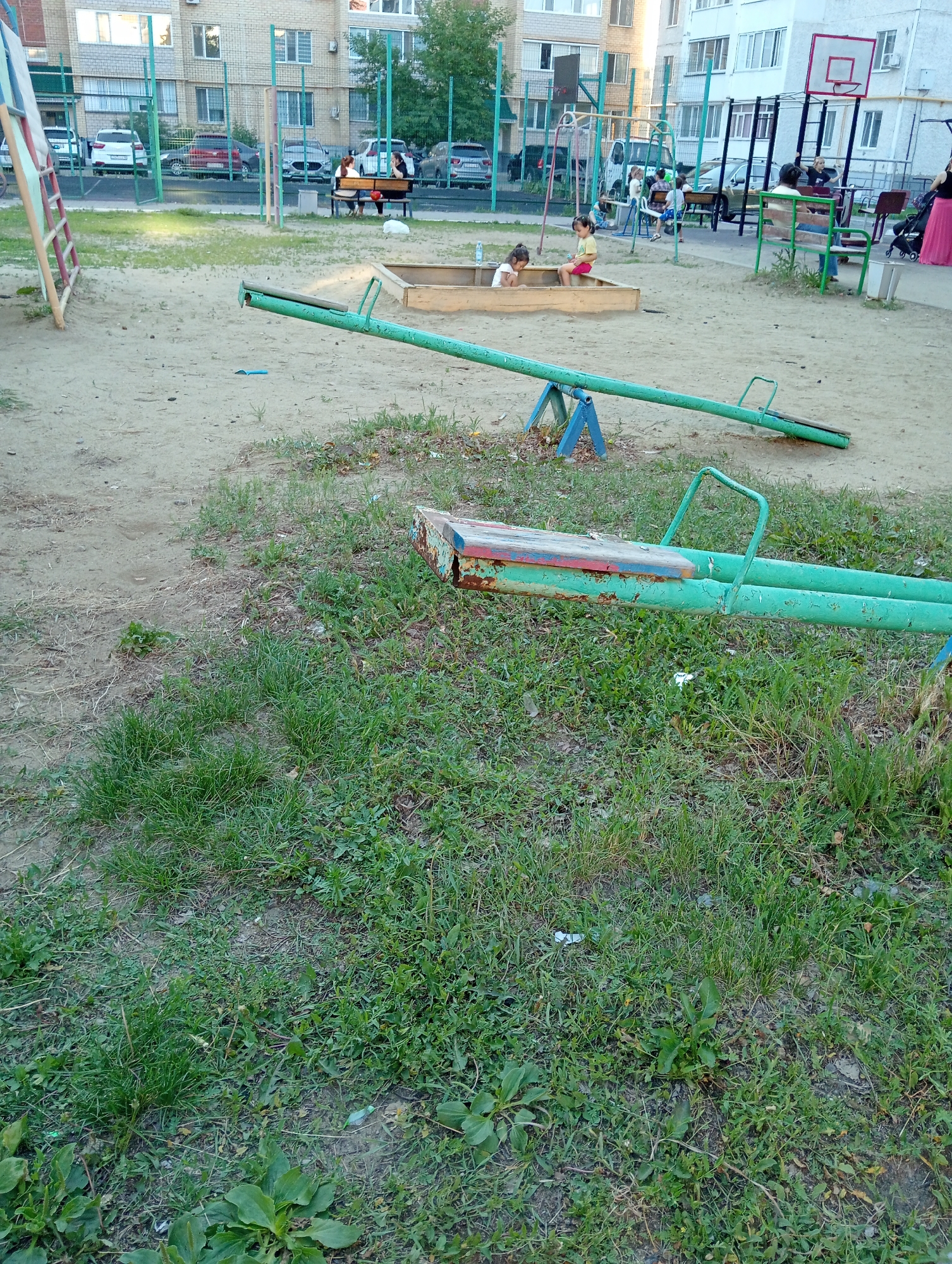 The image depicts an outdoor playground with a sandpit and a seesaw. In the background, there are apartment buildings and parked cars. Two young children, a girl in a yellow shirt and a girl with dark hair, are playing in the sandpit. There are other people in the background, some sitting on a bench and others standing. A woman in a pink skirt is also visible. The playground appears to be located in a residential area.