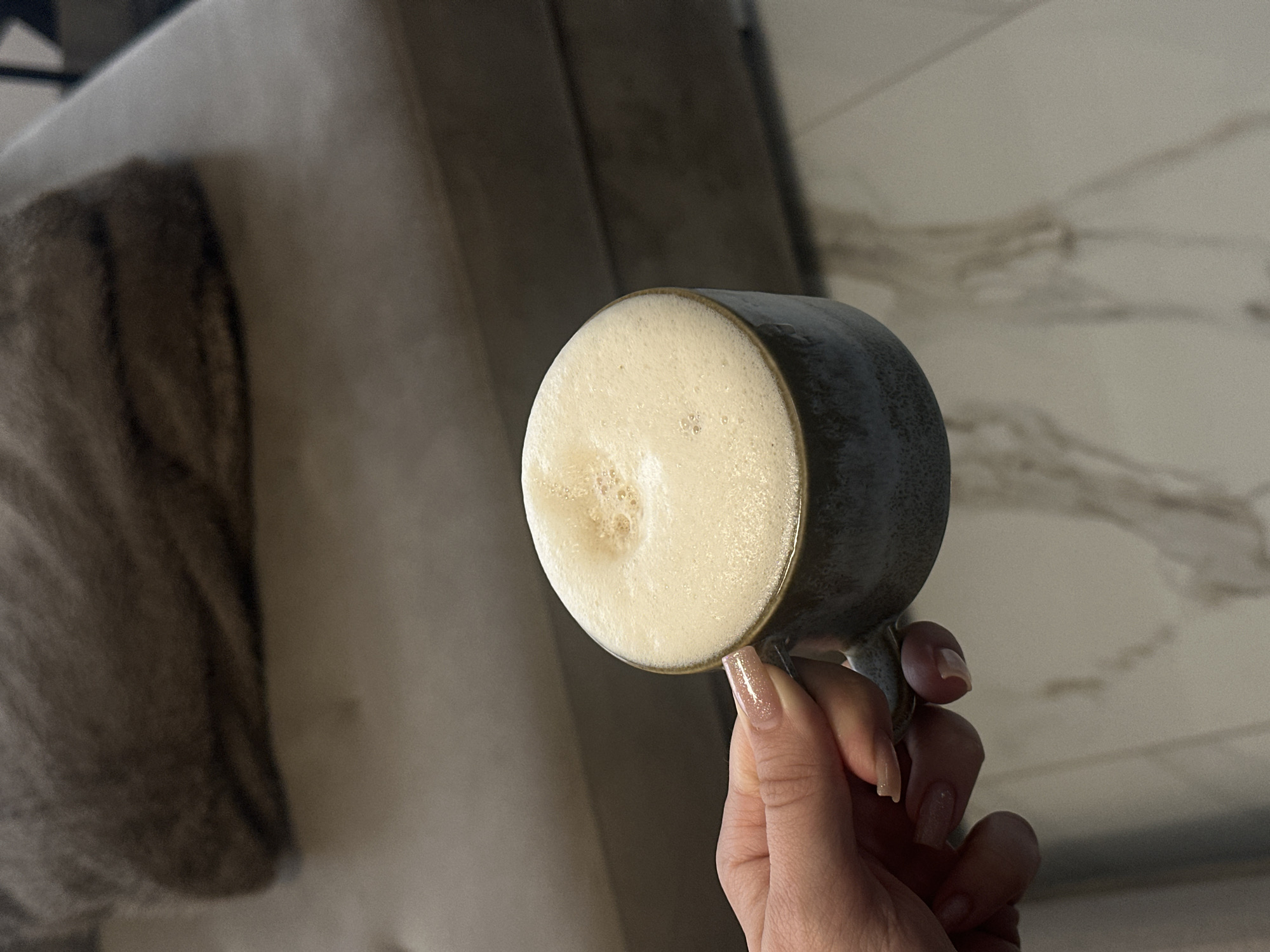 The image captures a close-up, angled view of a person's hand holding a mug filled with a frothy beverage. The main subject is a dark grey or muted blue ceramic mug, which holds a light brown drink topped with a thick, creamy layer of foam, characteristic of coffee or a similar hot beverage. Small and larger bubbles are visible on the foam's surface.
A hand, presumably female, with light skin and well-maintained, moderately long nails, grips the mug's handle. The nails are adorned with a light-colored, possibly iridescent or glittery polish.
The background is softly blurred, indicating a shallow depth of field. To the left, a portion of a light-colored, upholstered furniture item, such as a sofa or armchair, is visible, draped with a textured, patterned blanket or throw in shades of brown and grey. To the right, a lighter surface with subtle, marble-like patterns can be seen, likely a wall or a furniture surface. The overall setting suggests a comfortable indoor environment, possibly a living space.
The lighting is soft and diffuse, implying an indoor scene with ambient or artificial light. The activity depicted is the simple act of holding and enjoying a warm drink, conveying a sense of calm or relaxation. No text is visible within the image. The image itself provides no discernible cues to indicate its location as Sofia, Bulgaria.