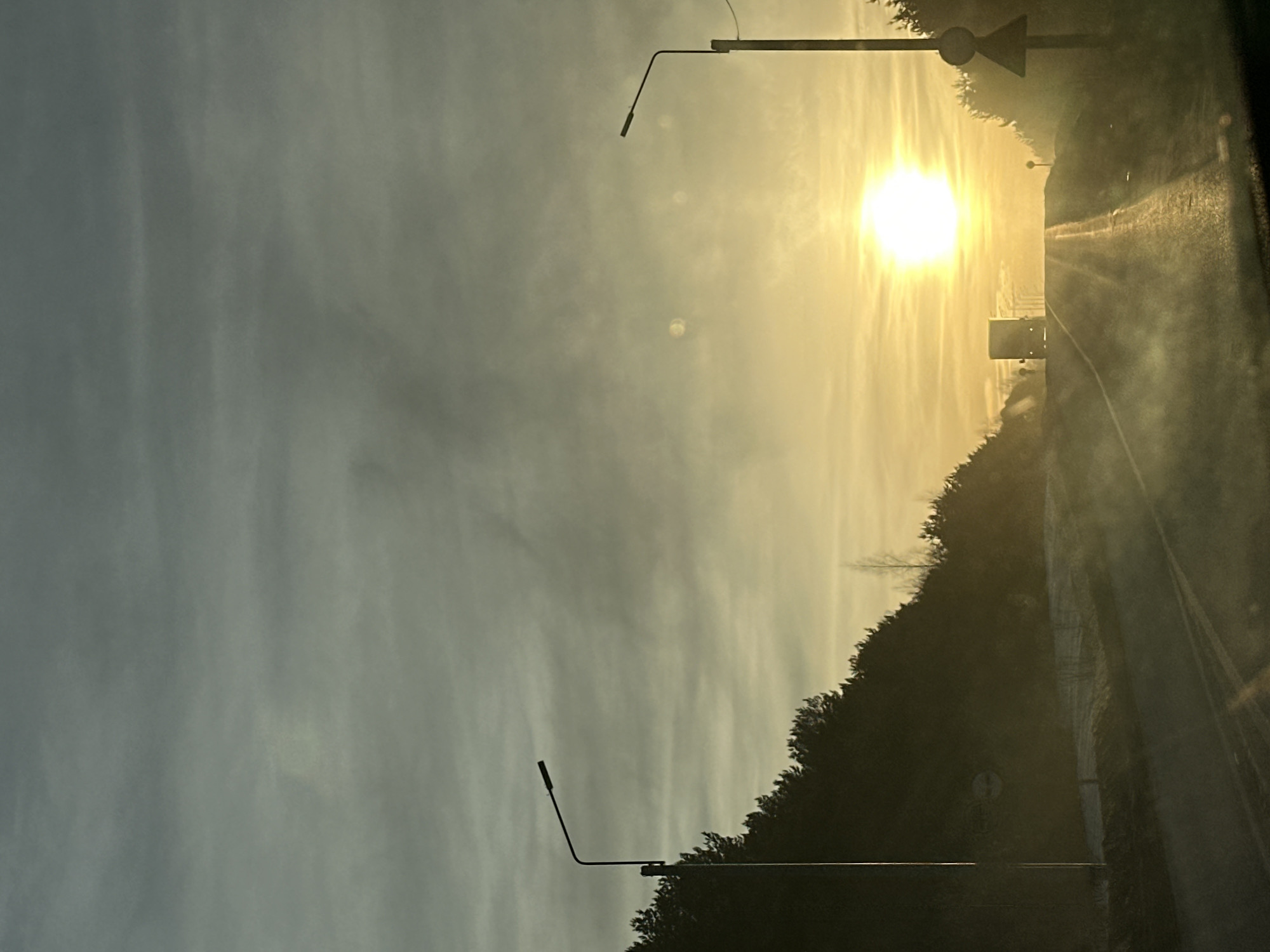 The image depicts a straight, two-lane road within Shud'ya, Russia, at either sunrise or sunset during winter conditions. The sun, a brilliant white orb, is positioned low on the horizon towards the center-right, casting an intense glare across the scene. The sky transitions from a muted grey-blue overhead to a strong yellow-orange directly above the horizon. Dense coniferous trees, appearing as dark silhouettes, line both sides of the road. Patches of snow are visible on the ground adjacent to the road. A dark street light pole with a single luminaire extends from the left side of the frame, with a circular road sign partially visible below it. Another dark street light pole is present on the right, alongside a triangular warning sign with a red border. A large, dark truck is observed in the distance, traveling away from the viewer, silhouetted against the bright sun as it ascends the road. Low-lying mist or haze is present over the road surface in the foreground, partially obscuring immediate ground details. No individuals are visible.