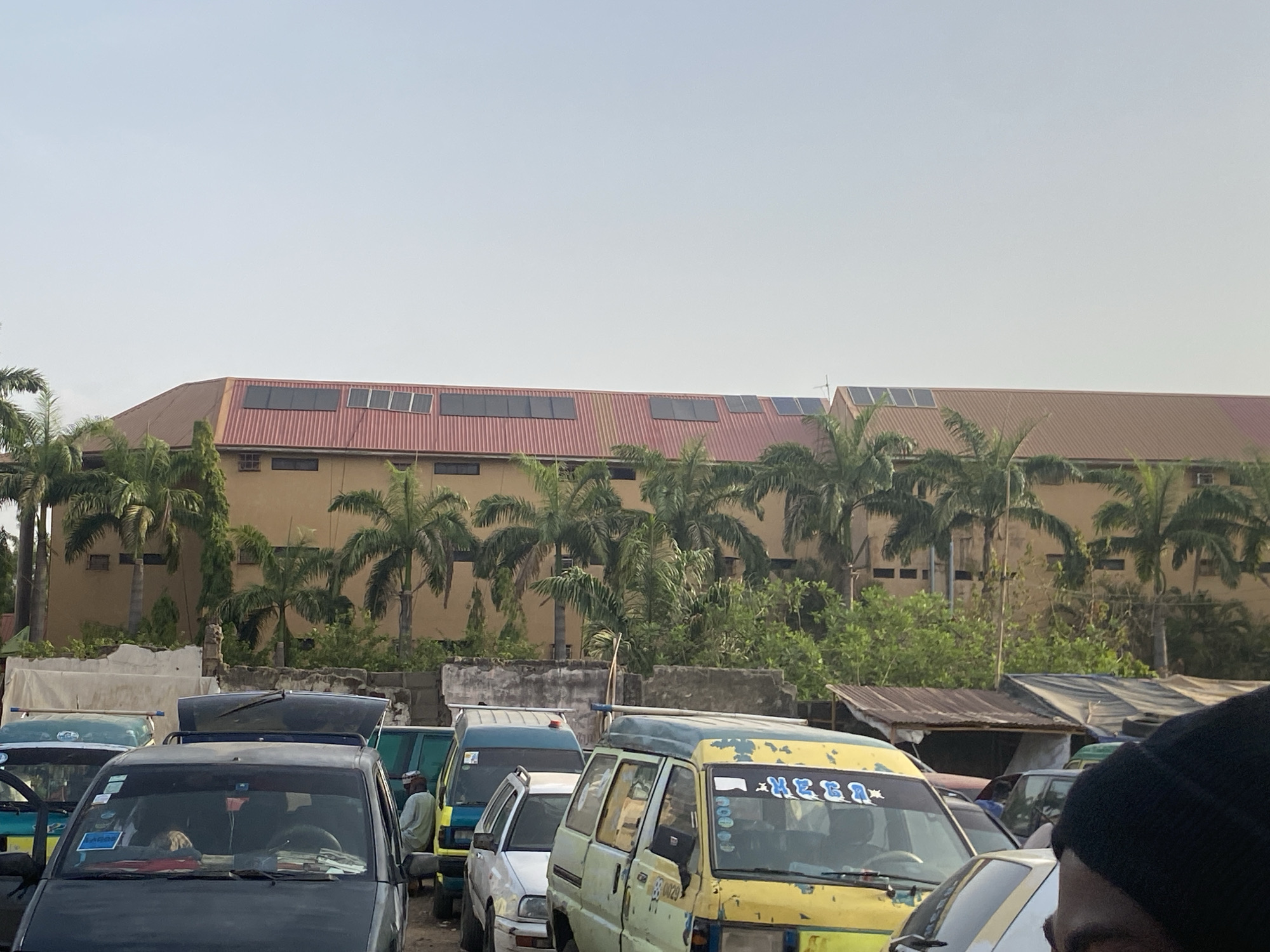 The image depicts a scene in Maiburiji, Nigeria, characterized by several commercial vehicles parked in an open area. A large, tan-colored building with a red roof is visible in the background, with solar panels installed on the roof. Palm trees are interspersed between the building and the vehicles, adding to the tropical ambiance. The vehicles appear to be public transportation vans and cars, with visible wear and tear on some. A man wearing a white robe can be seen standing near the vehicles. The sky is a pale blue, suggesting it could be early morning or late afternoon. The scene appears to be a transportation hub or parking area typical of an urban environment in Nigeria.