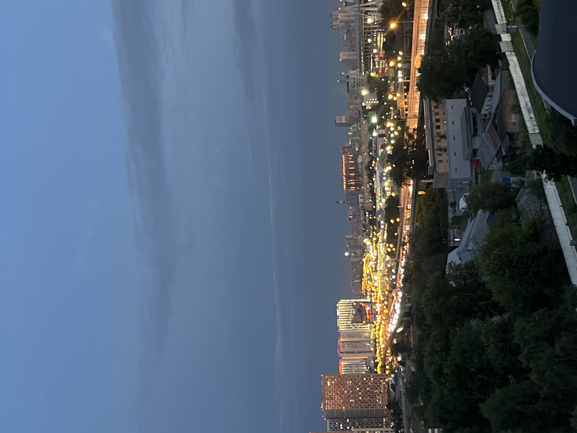 This image depicts an aerial view of a city at dusk or night. In the foreground, there is a dense cluster of dark green trees. Beyond the trees, a network of roads with numerous illuminated vehicles suggests active traffic. Several buildings are visible in the middle ground, including apartment complexes with illuminated windows and commercial structures. In the background, a cityscape with taller buildings and illuminated structures stretches towards the horizon. The sky is a deep blue with some wispy clouds. The overall scene conveys a sense of urban activity and illumination under a twilight sky, likely in Tarasovka, Russia, given the provided context. There are no people or specific actions clearly discernible in the image.
