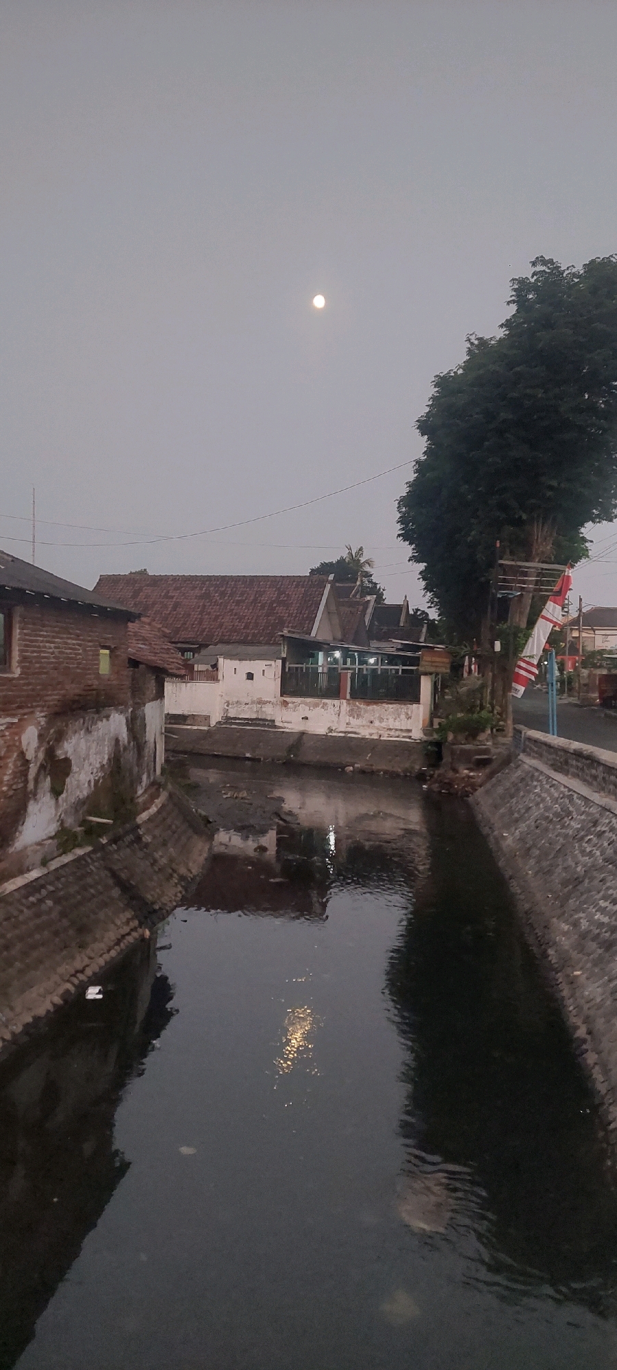 The image depicts a narrow canal or river flowing through a residential area in Lumajang, Indonesia, during what appears to be dusk or early morning given the soft, diffused light and the presence of the moon in the sky. The canal is lined with stone walls, and on either side, there are buildings with traditional tiled roofs.
On the left bank, a brick building with weathered walls stands prominently, with a single lit window suggesting occupancy. Across the canal, a cluster of buildings with white facades and terracotta roofs is visible. Some of these buildings appear to be shops or small businesses, with lights illuminated within. A tall, leafy tree with dense foliage is situated on the right bank, partially obscuring the view of the houses behind it. Indonesian flags, red and white, are attached to a pole near the tree.
The water in the canal is dark and still, reflecting the overcast sky and some scattered lights from the buildings. The overall scene conveys a quiet, possibly rural or semi-urban atmosphere, characteristic of many Indonesian towns. The presence of the moon suggests it is nighttime or very early morning.