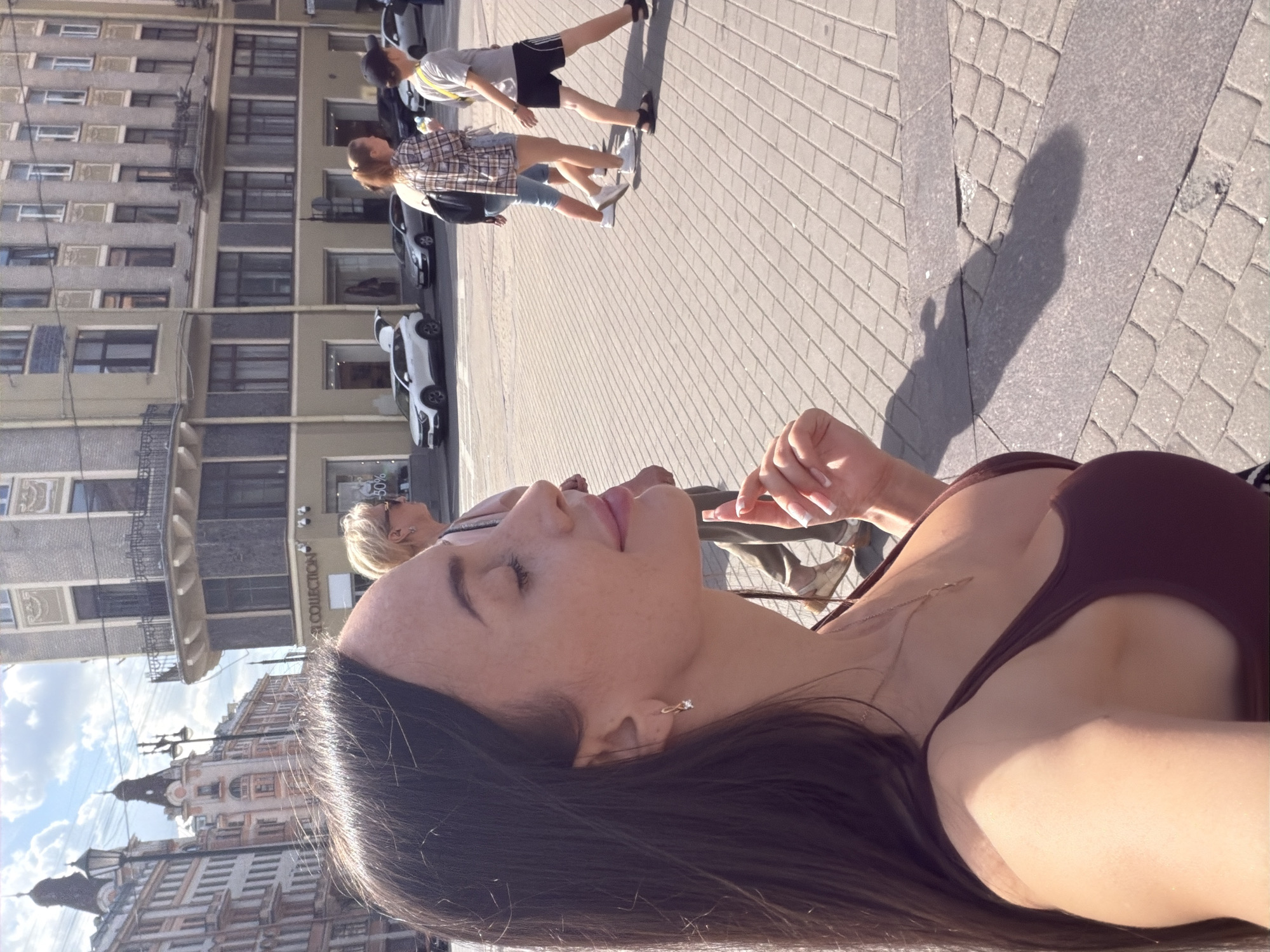 The image shows a woman in the foreground, looking to the right with her eyes closed and a slight smile. She has long, dark hair, is wearing a dark red tank top, and a delicate necklace. Her right hand is raised, fingers splayed. In the background, several people are walking on a paved street lined with buildings. The architecture suggests a European city, and the text on the building in the middle ground, "COLLECTION," along with the overall style, points to Saint Petersburg, Russia. Specifically, this scene appears to be on Nevsky Prospekt, a major avenue in Saint Petersburg, known for its historic buildings and pedestrian traffic. The day is sunny, casting shadows on the pavement.