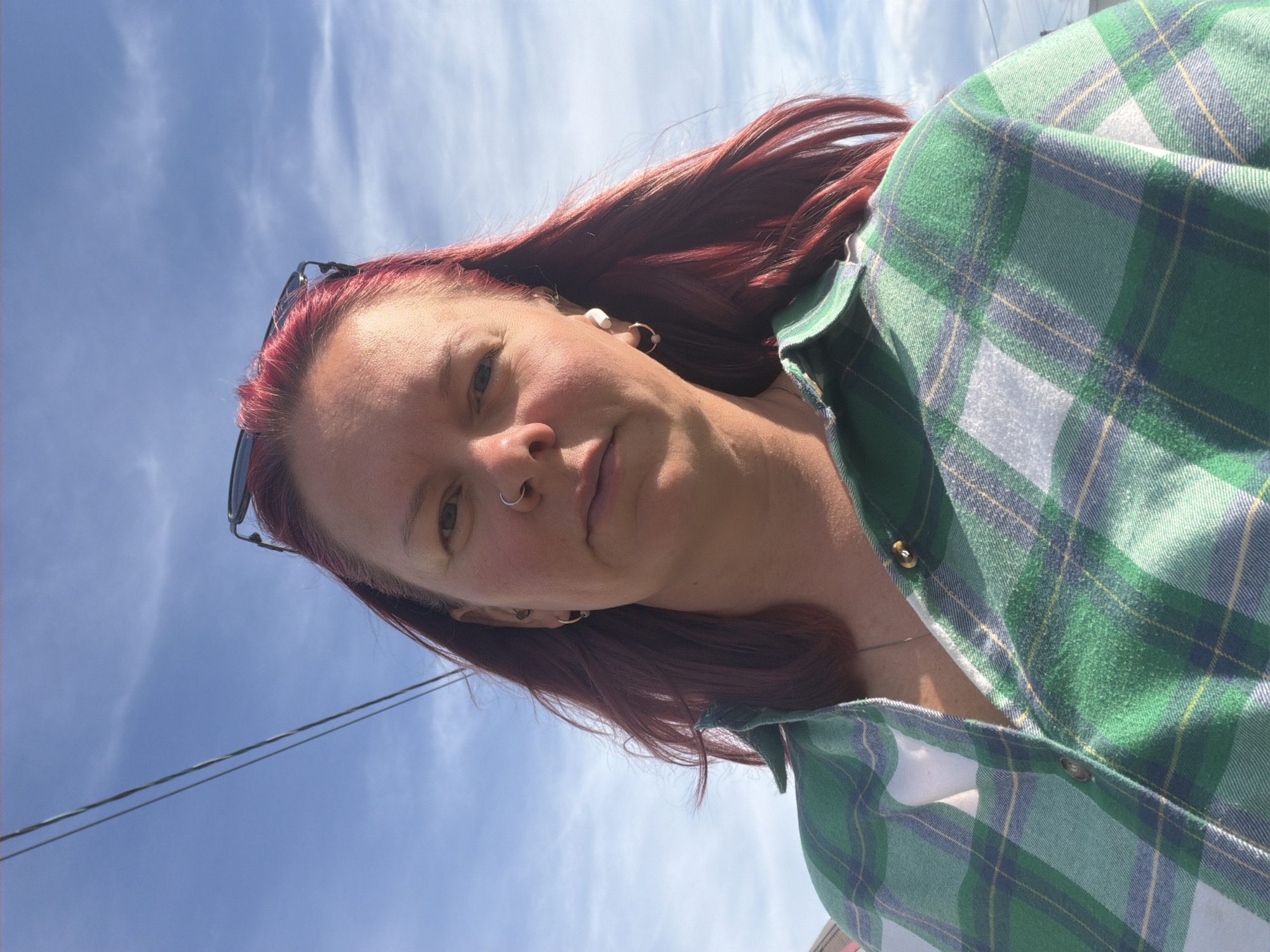A selfie taken outdoors. The subject is a woman with red hair, a nose ring, and sunglasses perched on her head. She wears a green and white plaid shirt. The background features a blue sky with wispy clouds. Two overhead cables are visible in the top left corner. The location is not discernible from the image.