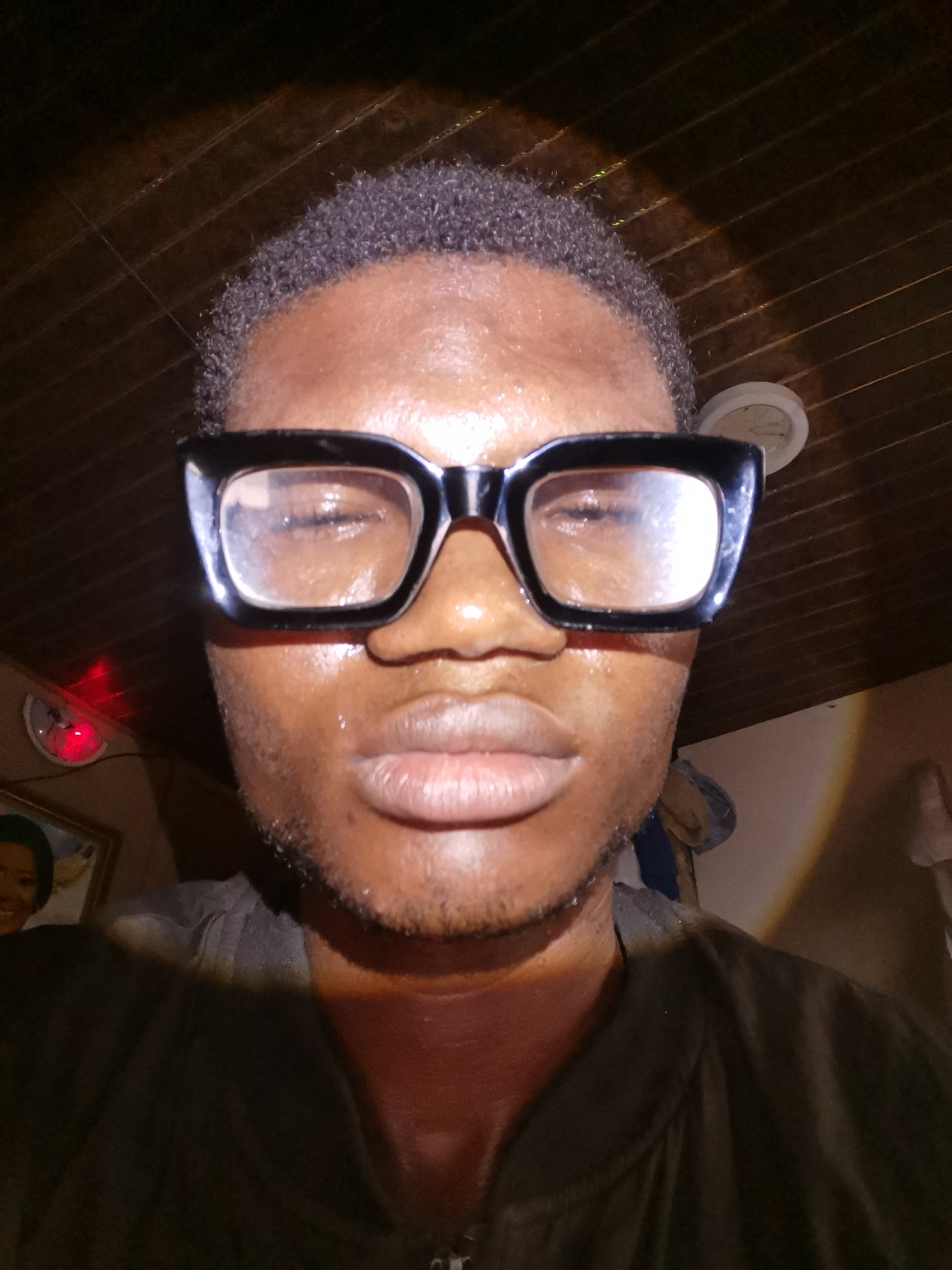 A close-up selfie depicts a man with dark skin wearing large, black-framed eyeglasses. His eyes are partially closed. He has short, dark hair. The room has a dark, wooden-planked ceiling. A ceiling fan is visible. On the left, a clock with a red light and a framed picture are partially visible. The man is wearing a dark shirt or jacket. The location is consistent with an interior in Lagos, Nigeria based on the environment.