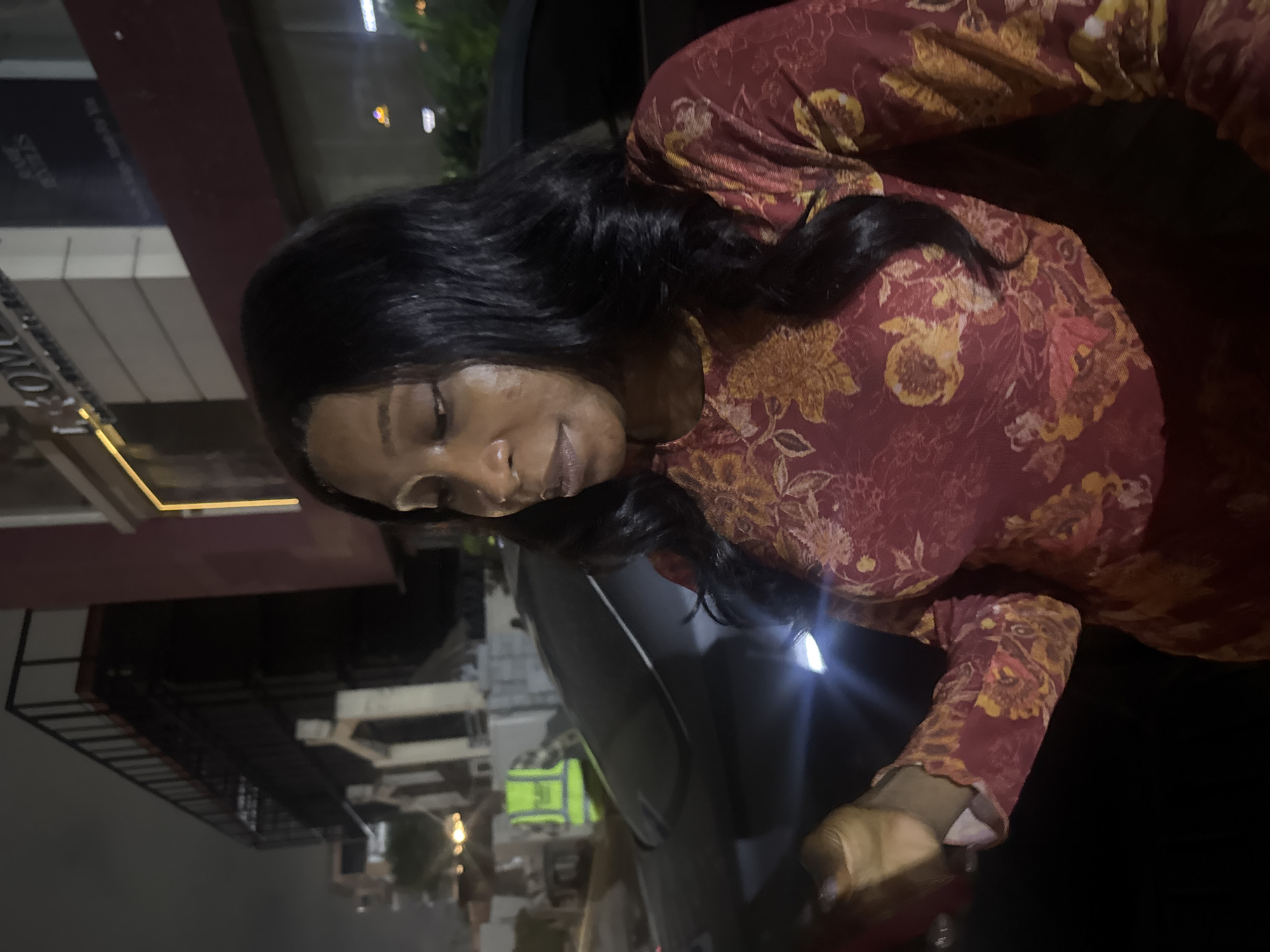 In Abuja, Nigeria, at night, a woman with dark hair stands in front of a car. She is wearing a long-sleeved red shirt with a floral pattern. The background shows a building with a dark facade and illuminated windows, suggesting a commercial area or hotel. The lighting is dim, typical of nighttime city environments. A person in a high-visibility vest is visible in the background, possibly a security guard or worker. The overall scene appears to be an urban street or parking area.