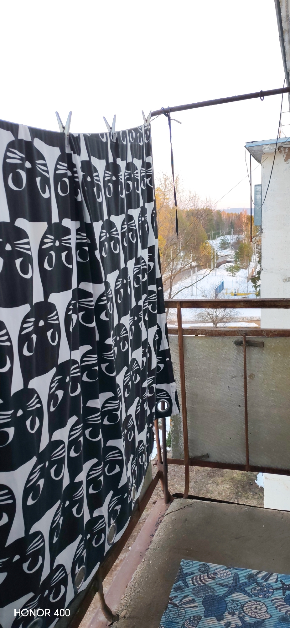 The scene captures a view from a balcony in Noviy, Russia. A black and white fabric curtain, patterned with repeating stylized cat faces, hangs from a rusty metal bar in the left foreground, secured by several white clothespins. The balcony's floor consists of weathered concrete, partially covered by a blue mat featuring seashell and starfish designs. A rusted metal railing, incorporating a translucent panel, frames the lower portion of the outdoor view. Beyond the balcony, a winter or early spring landscape is visible, characterized by snow-covered ground, bare deciduous trees, and scattered evergreens. A snowy path or road meanders through the terrain, with utility poles or similar structures present in the distance. An adjacent building with a weathered, light-colored facade and visible electrical wiring is partially observed on the right side of the frame. The sky is pale, indicating diffused light conditions.