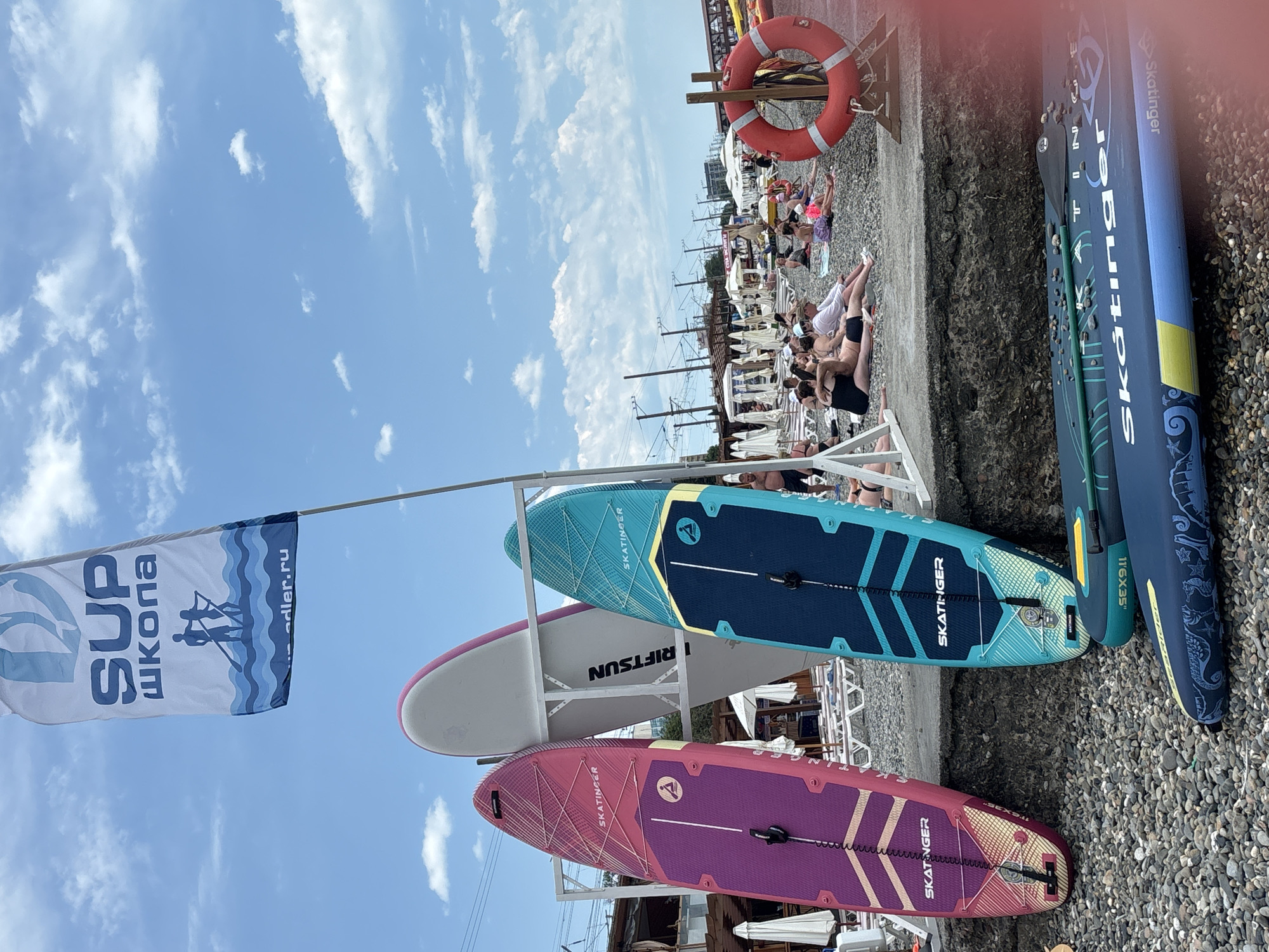 This media file captures a sunny day at a beach in Sochi, Russia, with a clear blue sky dotted with fluffy white clouds. The scene is a bustling resort area with many people relaxing on lounge chairs and sunbathing on the pebble beach. In the foreground, several stand-up paddleboards (SUPs) are displayed, leaning against each other or propped up, indicating a potential watersports activity or rental service.
A prominent flag in the lower left corner reads "SUP ШКОЛА" (SUP School) in large blue letters, with "adler.ru" visible below, suggesting a local surf school in Adler, a district of Sochi. The paddleboards themselves bear branding, including "SKATINGER" and model specifications like "11'6X35". One board also has "RIFTSUN" printed on it. A red life preserver ring is mounted on a wooden structure in the background, adding to the beachside atmosphere. The overall mood is one of leisure and recreation on a bright, warm day.