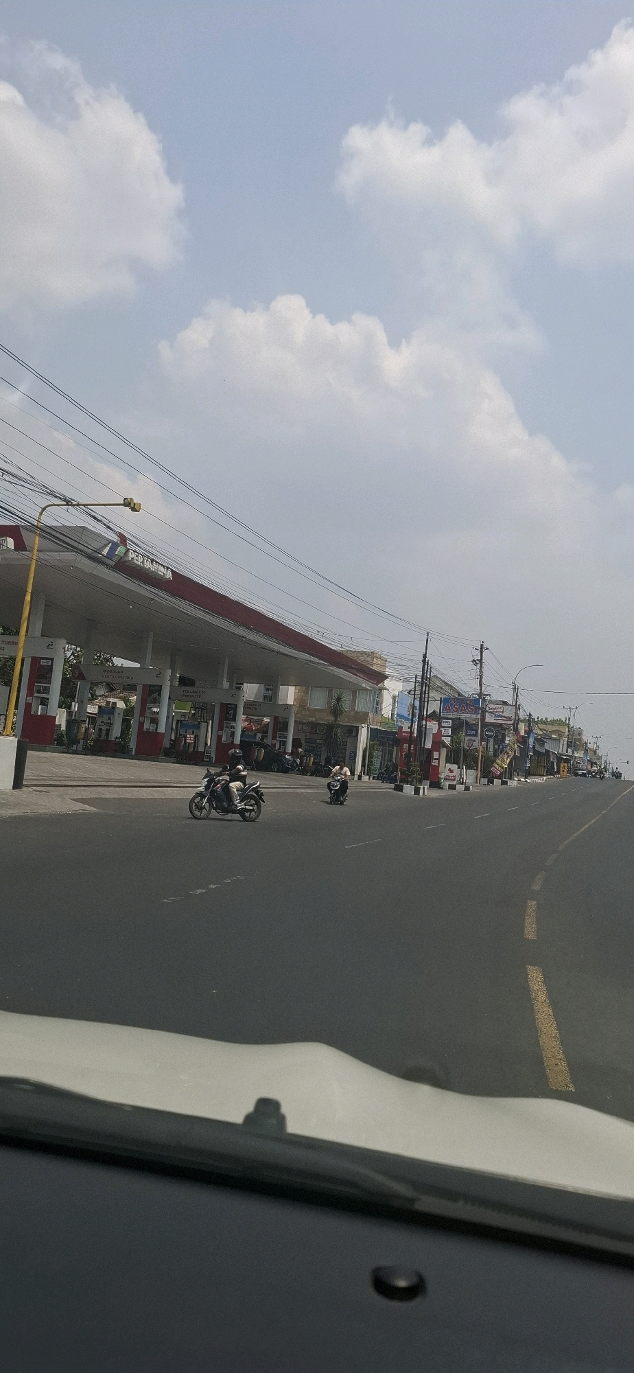 This image depicts a daytime scene on a paved road in Colomadu, Indonesia, with a Pertamina gas station visible on the left. The sky is partly cloudy with white cumulus clouds against a pale blue background. A yellow traffic signal is mounted on a pole above the road.
The Pertamina gas station features a red and white canopy covering multiple fuel pumps. Signs indicate various fuel types. Adjacent to the gas station are several buildings, some appearing to be shops or businesses, with signage including "ASAS" and other unidentifiable text. Electrical wires are strung across the scene, supported by poles.
Two motorcycles are present on the road. The first, in the foreground and slightly left of center, is ridden by a person wearing a helmet and dark clothing. The second motorcycle is further down the road, also with a rider. The road has yellow lane markings. In the bottom foreground, the edge of a white vehicle's dashboard and windshield is visible, suggesting the photo was taken from inside a car.