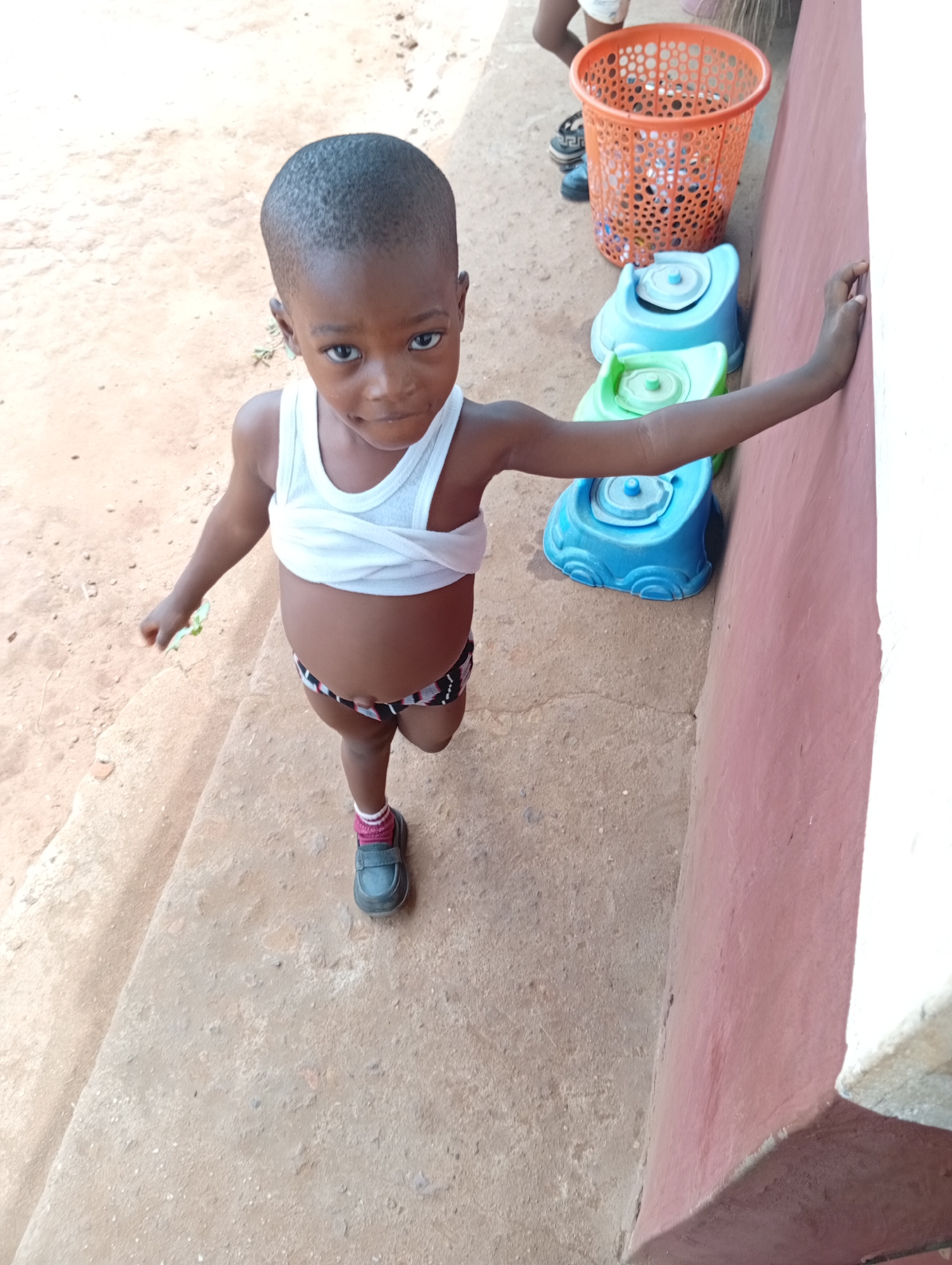 A young child stands near a light red and white wall in Ukwu-Abwa, Nigeria. The child wears a white tank top and patterned underwear, along with one sock and a single shoe. To the right of the child, plastic toy potties in shades of blue and green are lined up next to the wall, with an orange plastic basket filled with items visible above them. The ground is concrete.