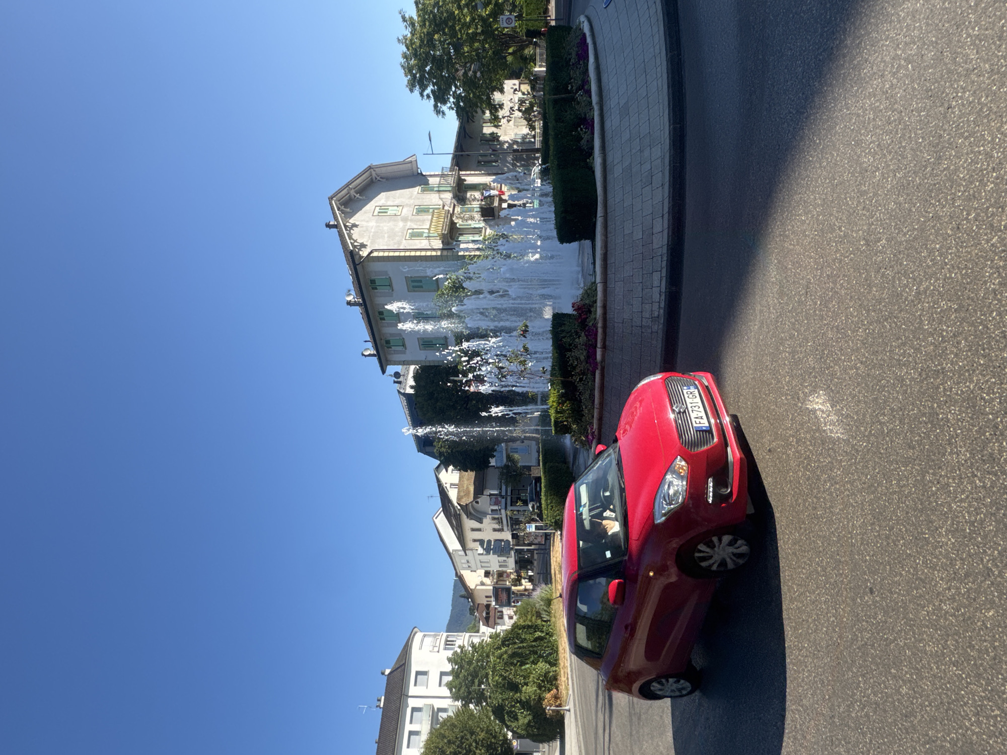 A red Suzuki Swift is turning through a roundabout in Divonne-les-Bains, France. A woman is visible behind the steering wheel. The roundabout features a large, multi-tiered fountain with water spraying upwards, surrounded by manicured green hedges and flowering plants. In the background, buildings with light-colored facades and green shutters line a street. Trees with lush green foliage are present, and the sky is clear and blue. The image is captured on a sunny day, indicated by the strong shadows cast on the road. The license plate of the car is visible and reads "FA 731 GR".