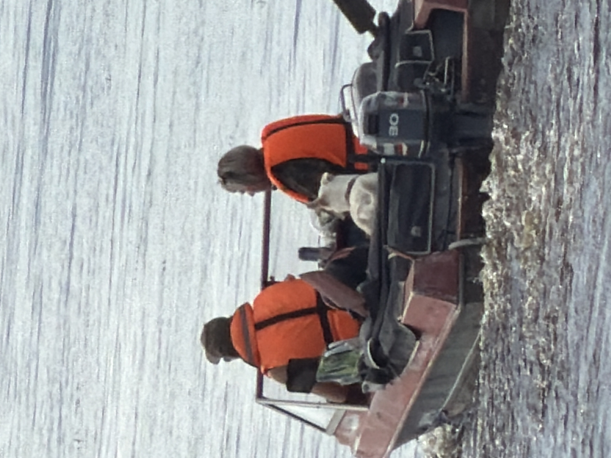 Two people and a dog are in a boat on a body of water. The person on the left is wearing an orange life vest and a hat, and is operating the boat. The person on the right is wearing an orange life vest and is looking to their left. A white dog is lying down between the two people. The boat is red and black and has a windshield. There is an outboard motor on the back of the boat. The water is choppy. The scene is likely taking place in the city of Solikamsk, Russia, as indicated by the context provided.