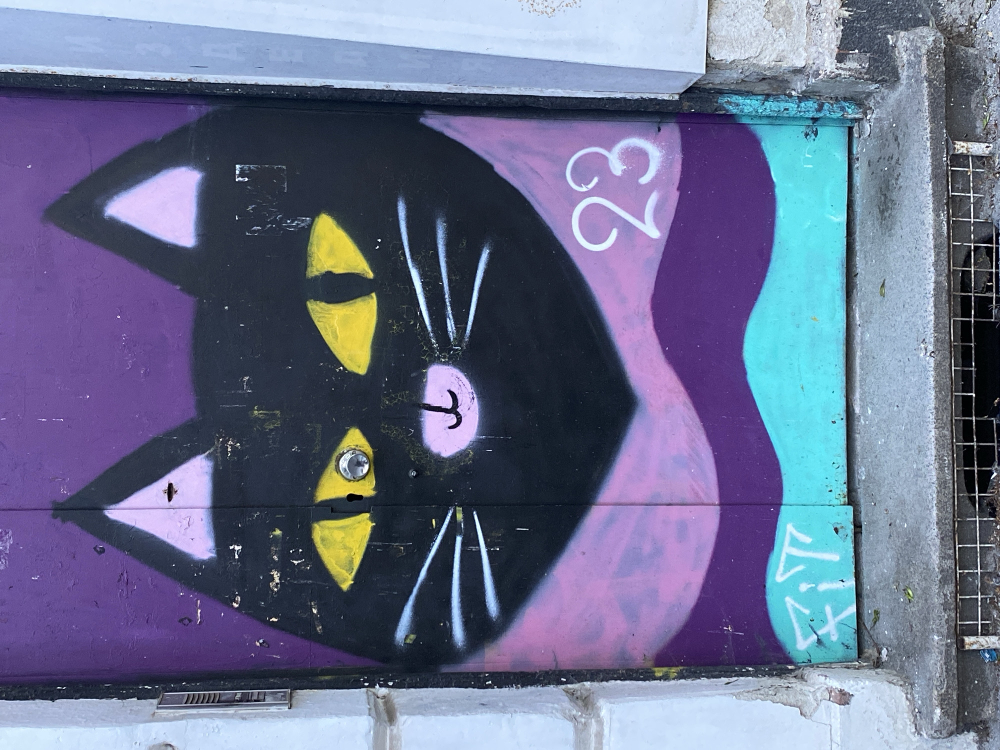 An outdoor, waist-level view features a section of graffiti-covered wooden door or covering, likely an entryway. The graffiti depicts a stylized black cat face with triangular pink inner ears, downward-pointing yellow eyes, white whiskers, and a pink nose. The background surrounding the cat is composed of purple, pink, and turquoise sections. The number "23" is painted in white on the purple and pink background, near the right side of the cat's head. A white graffiti signature "KIL" is visible in the lower right corner, painted on the turquoise section. The structure is bordered at the top by a white-painted surface and on the right by a concrete curb and a metal drainage grate. The image was taken in Sofia, Bulgaria.