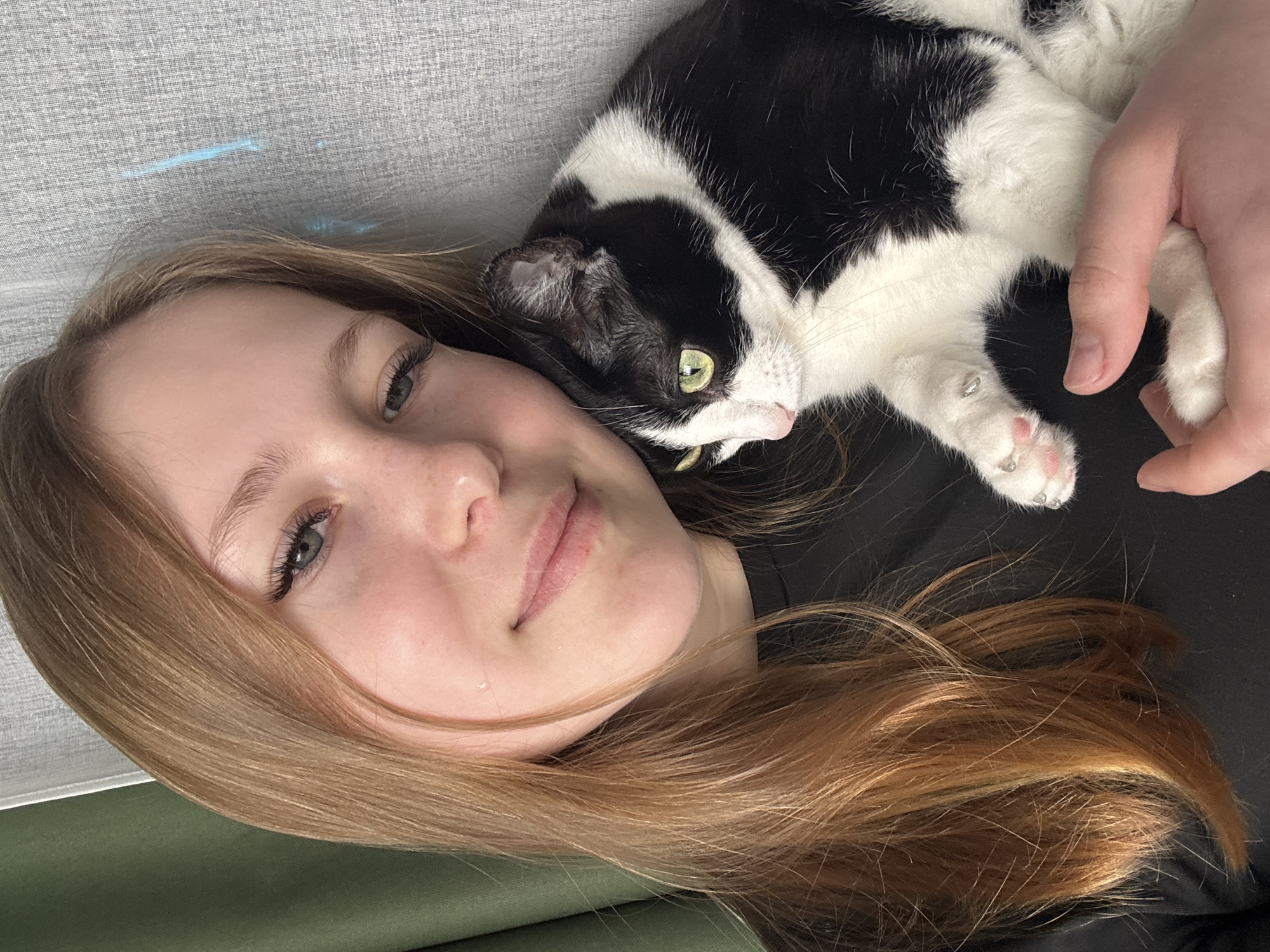 A young woman with light skin and long, light brown hair is lying down with a black and white cat. The woman is positioned on a gray textured surface. The cat is lying on or beside her, with its paws near her head. The woman has green eyes. A hand is touching the cat. A dark fabric covers the background. The photograph was taken indoors. It is not possible to determine the location as Chaikovsky, Russia from the image alone.