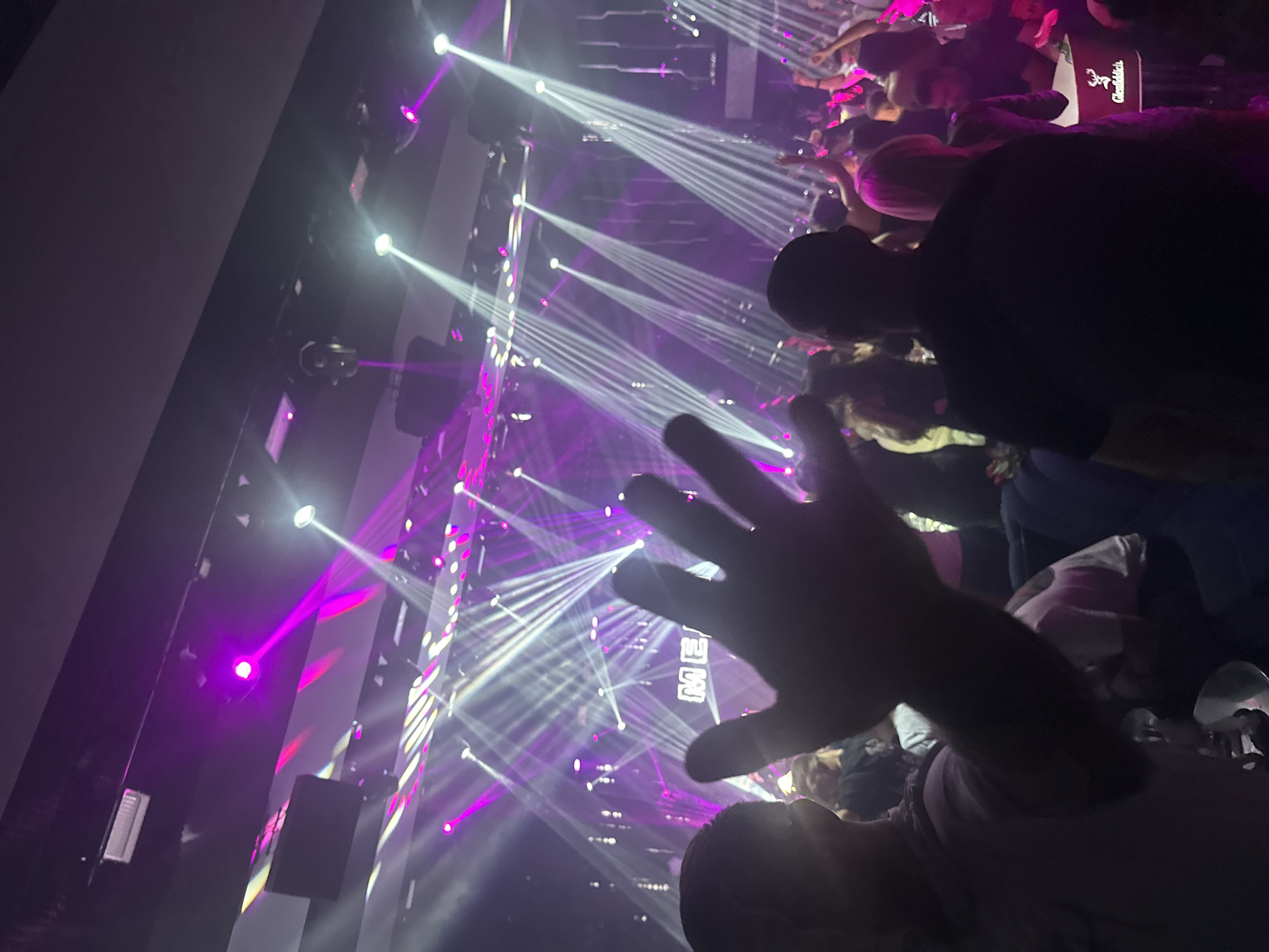 A dense crowd is gathered inside the Metropolis Club in Sliven, Bulgaria. Overhead, multiple lights are emitting intense beams of white and purple light, creating a dynamic, energetic atmosphere. In the foreground, a person holds up their hand, obscuring a portion of the view. Several other individuals are visible, their faces partially obscured by the low lighting and the density of the crowd. One person is wearing a black shirt and is partially turned away from the camera. The club appears to be packed.