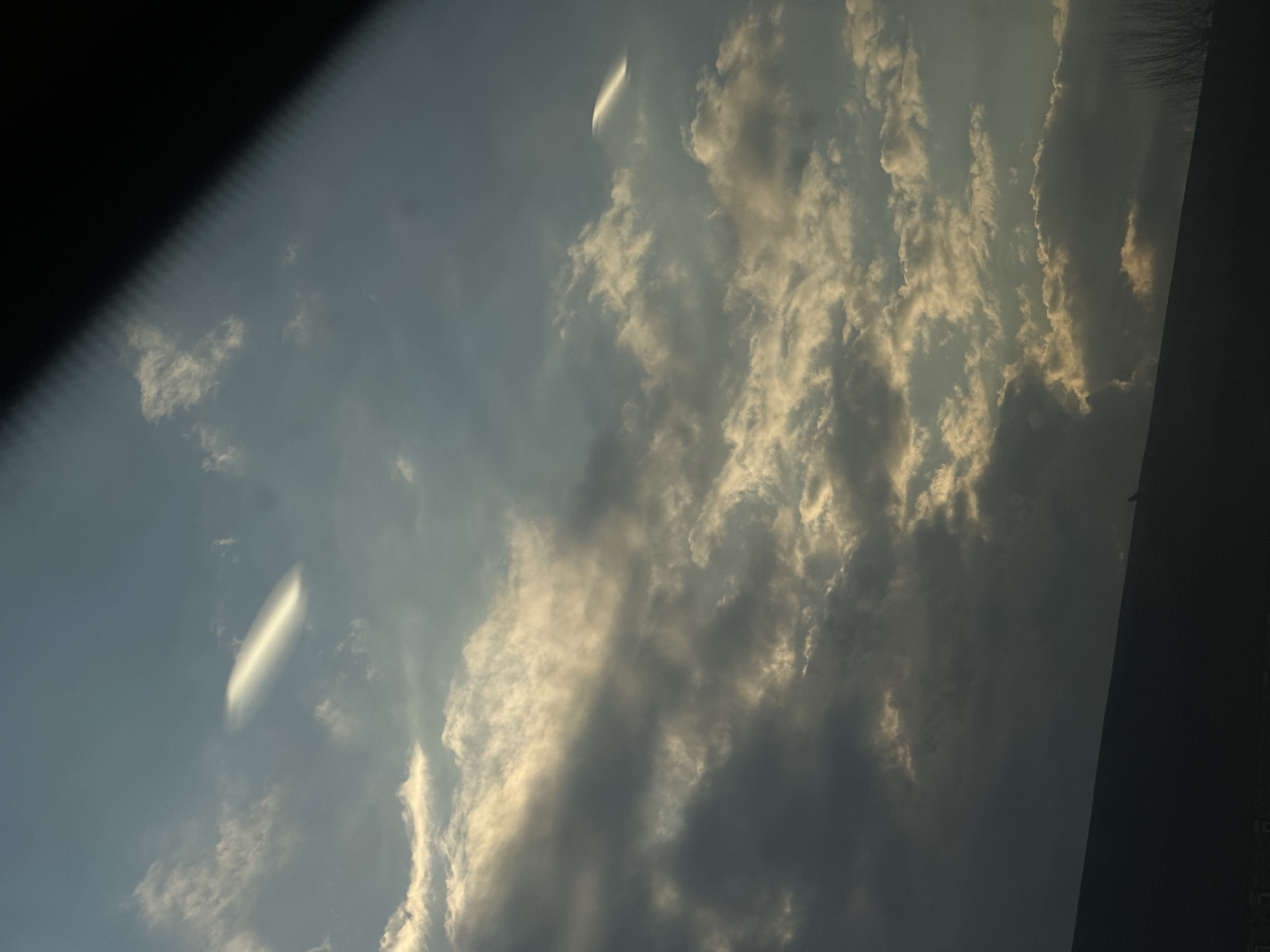 The image shows a sky filled with clouds, with some areas of blue visible. There are two distinct lenticular clouds that are bright white, appearing like elongated lenses. The overall lighting suggests either sunrise or sunset, as the clouds are illuminated with a warm tone. A portion of a dark structure is visible on the upper left and right corners, and it's not apparent the location is London, United Kingdom.