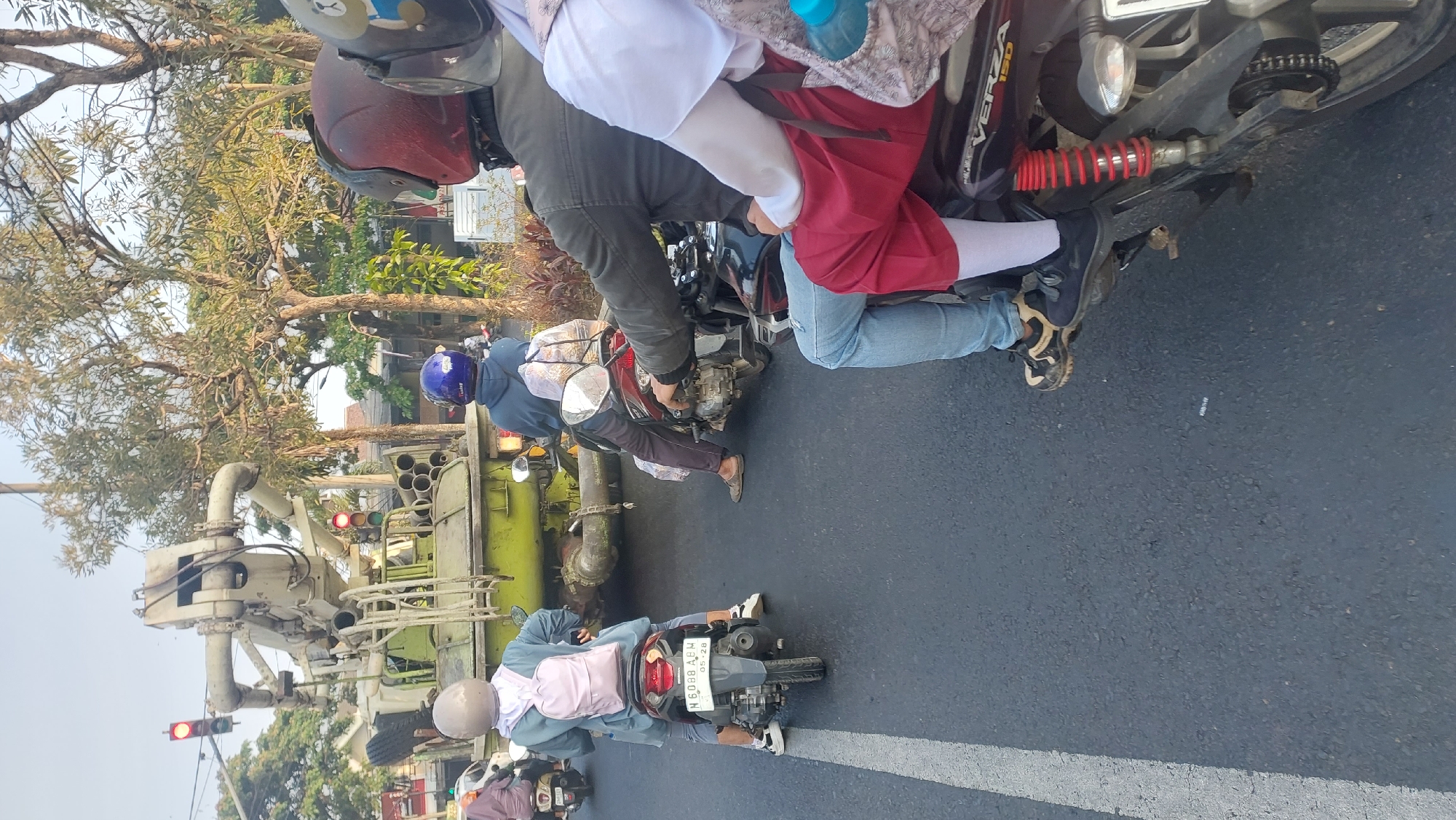 The image shows a street scene in Lesanpuro, Indonesia, with several motorbikes on a road. In the foreground, a motorbike is partially visible, with its rear wheel and suspension clearly depicted. Two individuals are seated on this motorbike; the passenger in the back wears a white hijab and a patterned fabric bag, while the rider in front wears blue jeans and dark shoes. To the left, another motorbike is seen from behind, carrying a person with a light-colored helmet and a pink backpack. Further back, a larger green vehicle, possibly a utility truck with a large boom or crane, is stationary. Several traffic lights are visible, with one showing red. Trees line the side of the road, and the overall lighting suggests it is daytime.