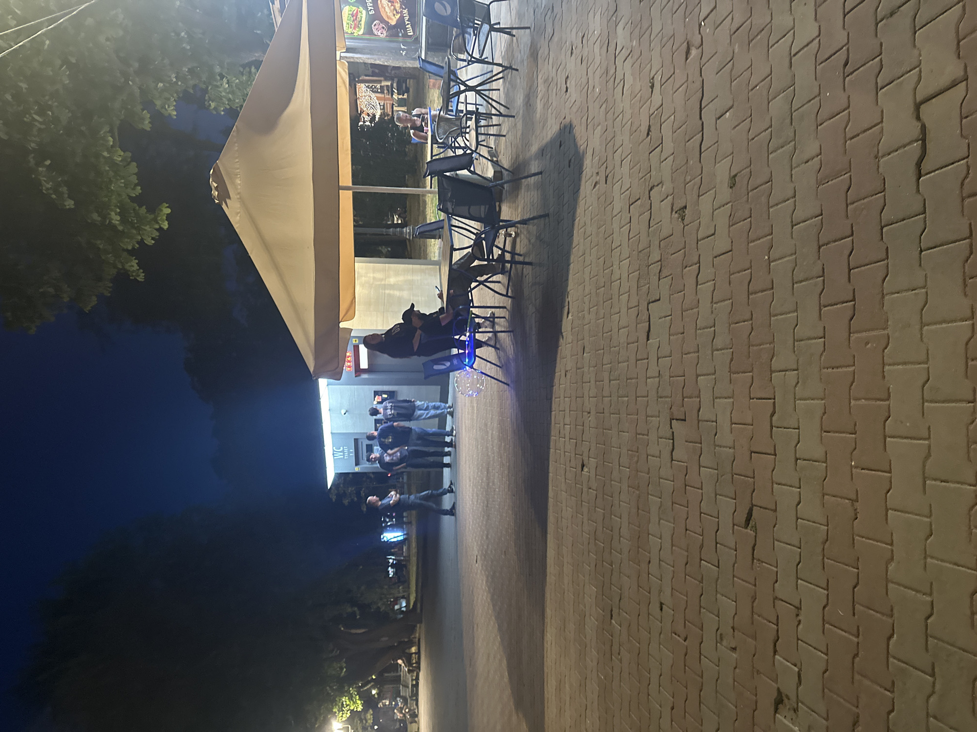 In the city of Odesa, Ukraine, a paved area in front of a cafe with outdoor seating is depicted at night. A light brown awning shades several tables and chairs. A few people are seated at the tables, one woman is looking at her phone. A food kiosk with a sign indicating it sells burgers and shawarma is visible in the background. Several men are standing near a public restroom with a "WC" sign. The area is lit by artificial lights.