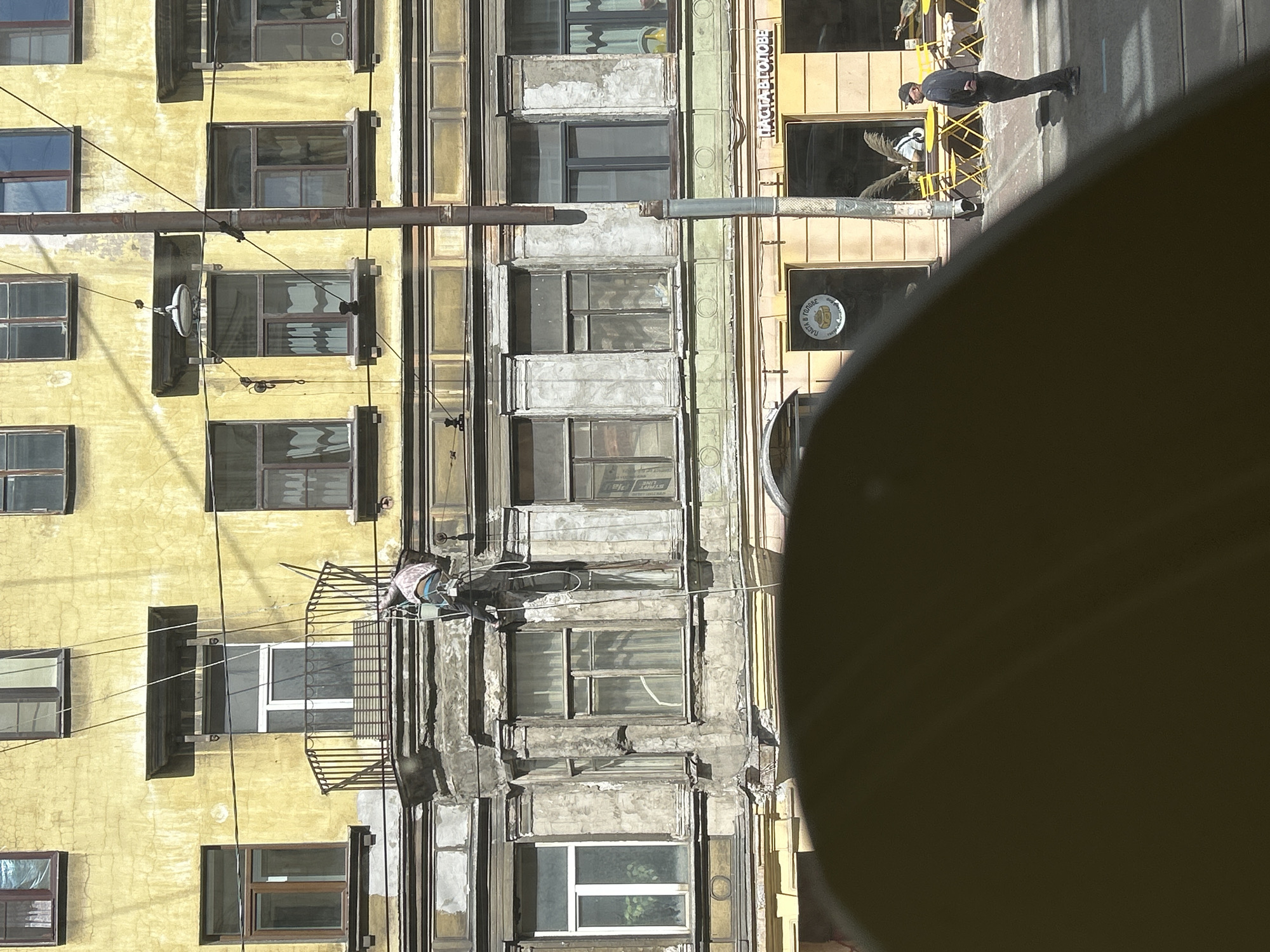 In a medium shot, a person is suspended by ropes on the side of a weathered yellow and grey building, seemingly engaged in construction or repair work. They are wearing a light-colored top and dark pants, with a bucket attached to their harness. The building features multiple windows, some with balconies, and ornate architectural details. In the foreground, a dark, out-of-focus shape dominates the bottom of the frame. To the right, a man in a dark jacket and baseball cap stands on the sidewalk looking towards the building, with yellow tables and chairs visible outside storefronts on the ground level. The scene is lit by bright sunlight, casting shadows.