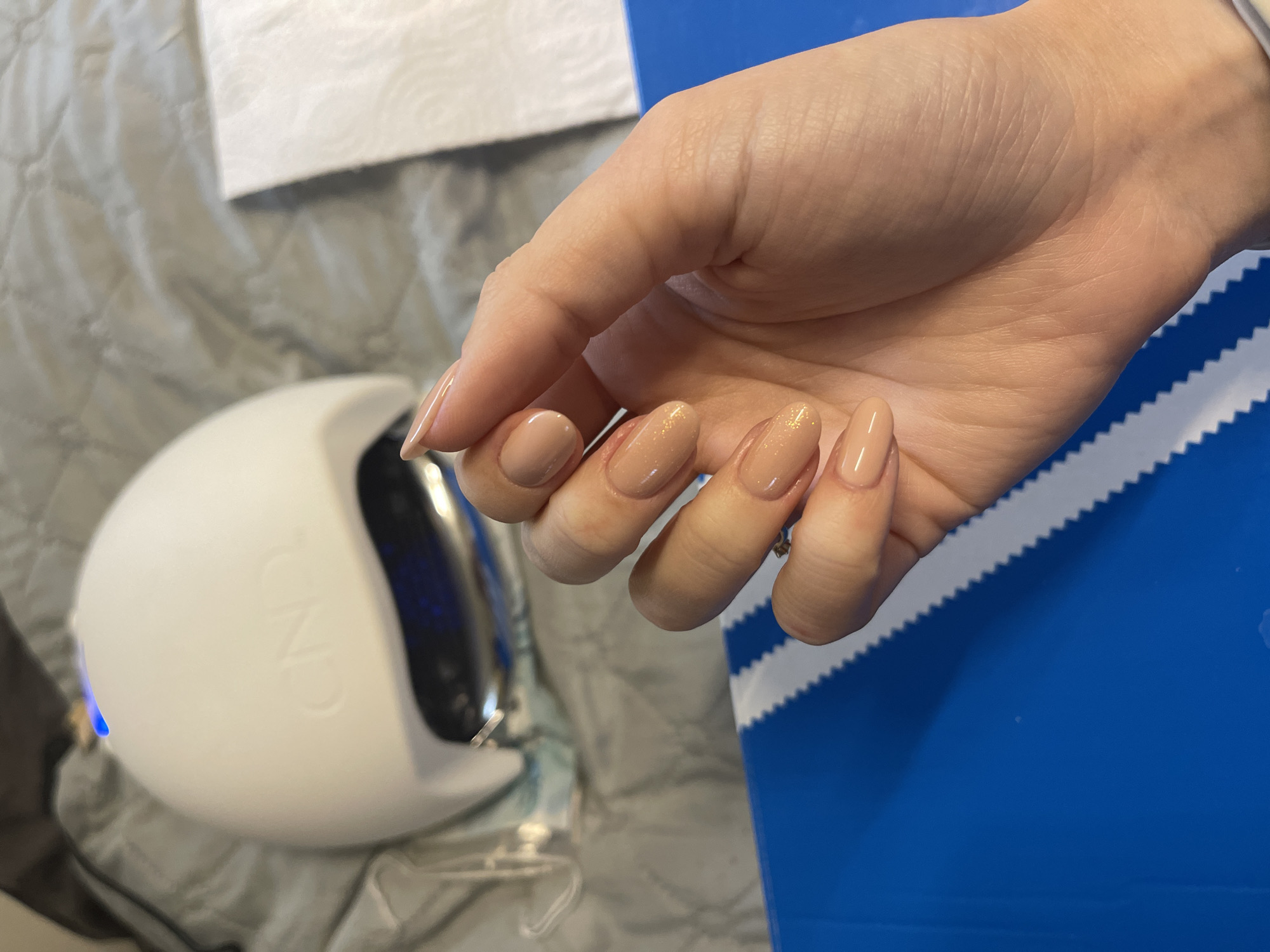 A close-up, overhead shot reveals a person's right hand with manicured nails, featuring a light beige/nude color, some with subtle gold glitter. The hand rests against a blue surface with white, serrated stripes, possibly an Adidas box. Adjacent to the hand, on the left, is a white CND nail lamp. A white paper towel is situated in the upper left corner. The backdrop appears to be a grey patterned surface, possibly a bedspread. The location is not discernible as being in Sofia, Bulgaria based on the visual information available.